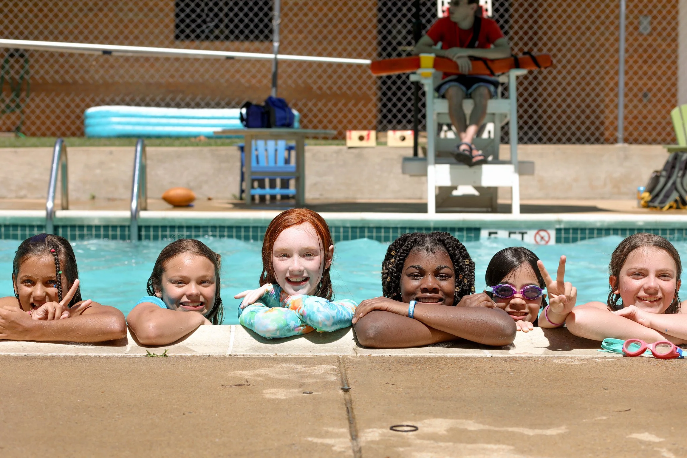 Six children smiling and leaning on the edge of a swimming pool, with a lifeguard sitting in a chair in the background, all enjoying a sunny day at the pool.