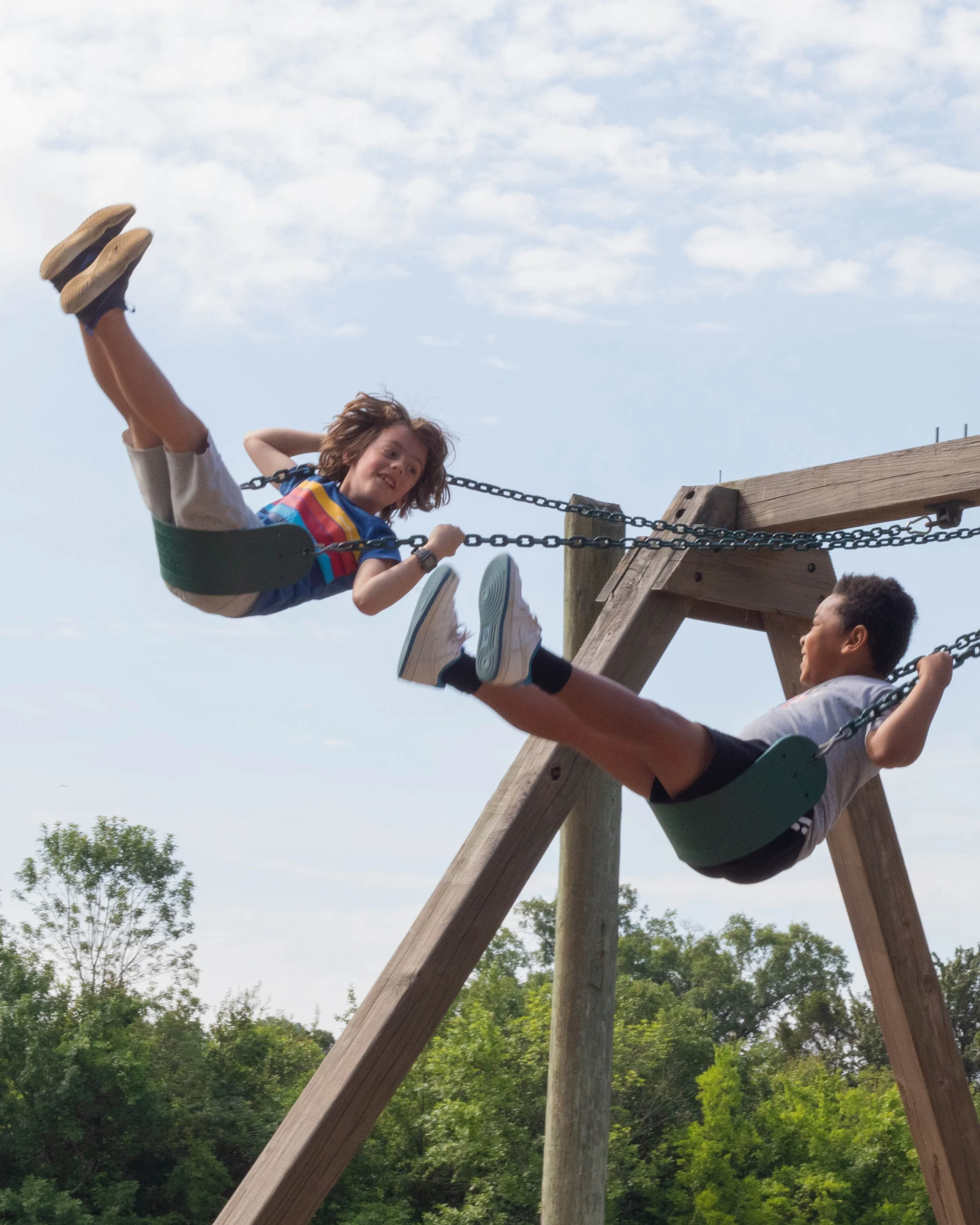 Two children, a girl and a boy, enjoying a swing set outdoors with a blue sky and trees in the background.