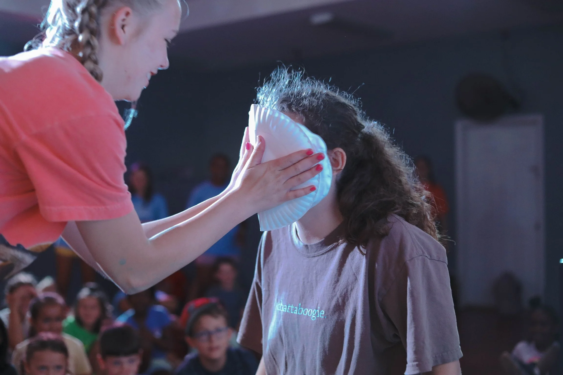 A woman is placing a paper plate over a girl's face in front of an audience, with the girl wearing a brown t-shirt and curly hair. The woman holds the plate with both hands and the girl is standing still. The scene appears to be indoors with several children and adults watching.