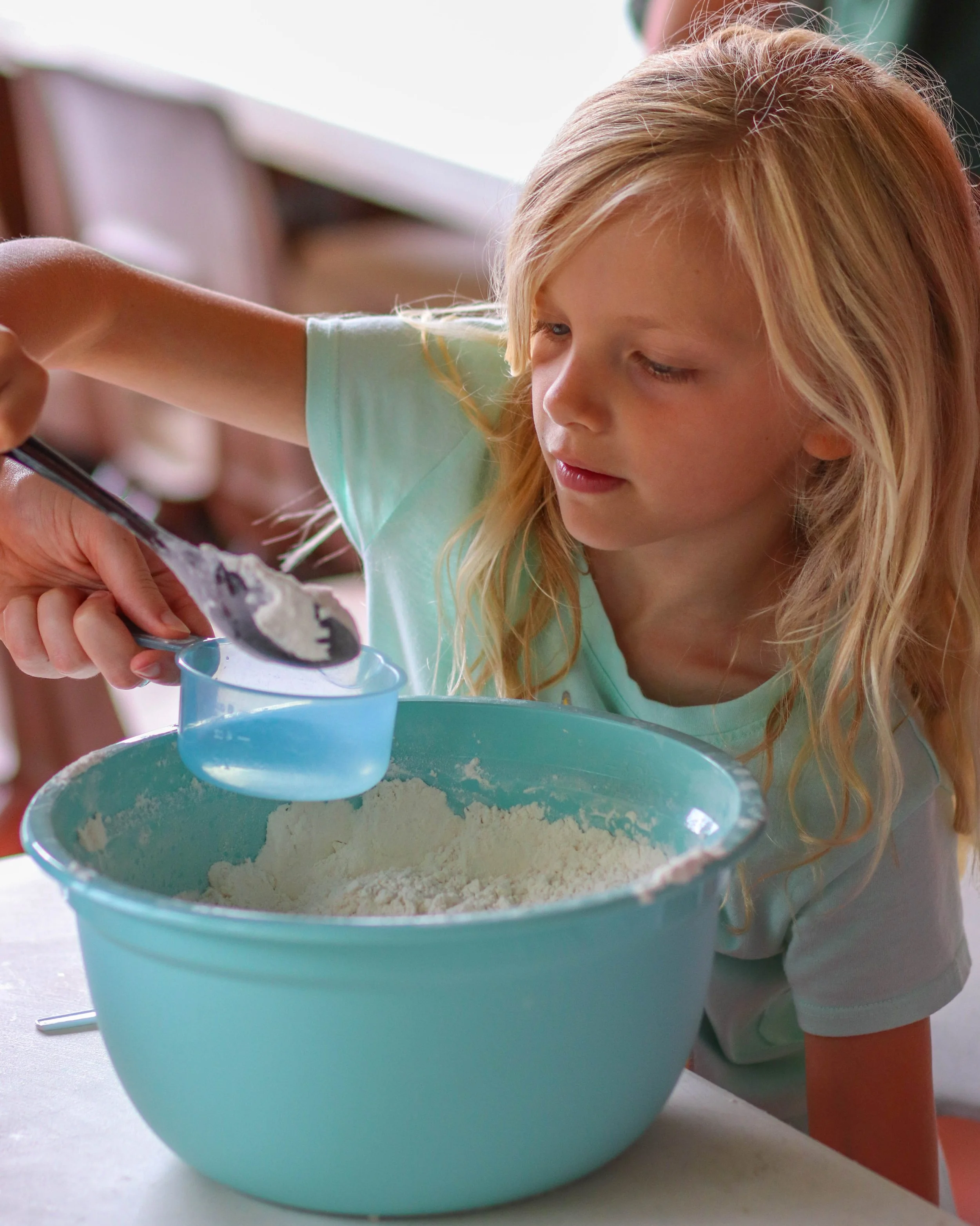 A young girl with blonde hair is using a spoon to measure flour into a blue mixing bowl.