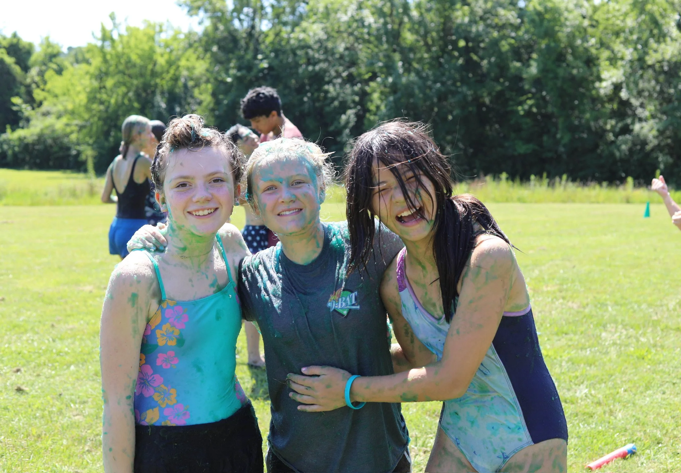 Three girls smiling and covered in colorful chalk or paint outdoors on a sunny day, with other children playing in the background.