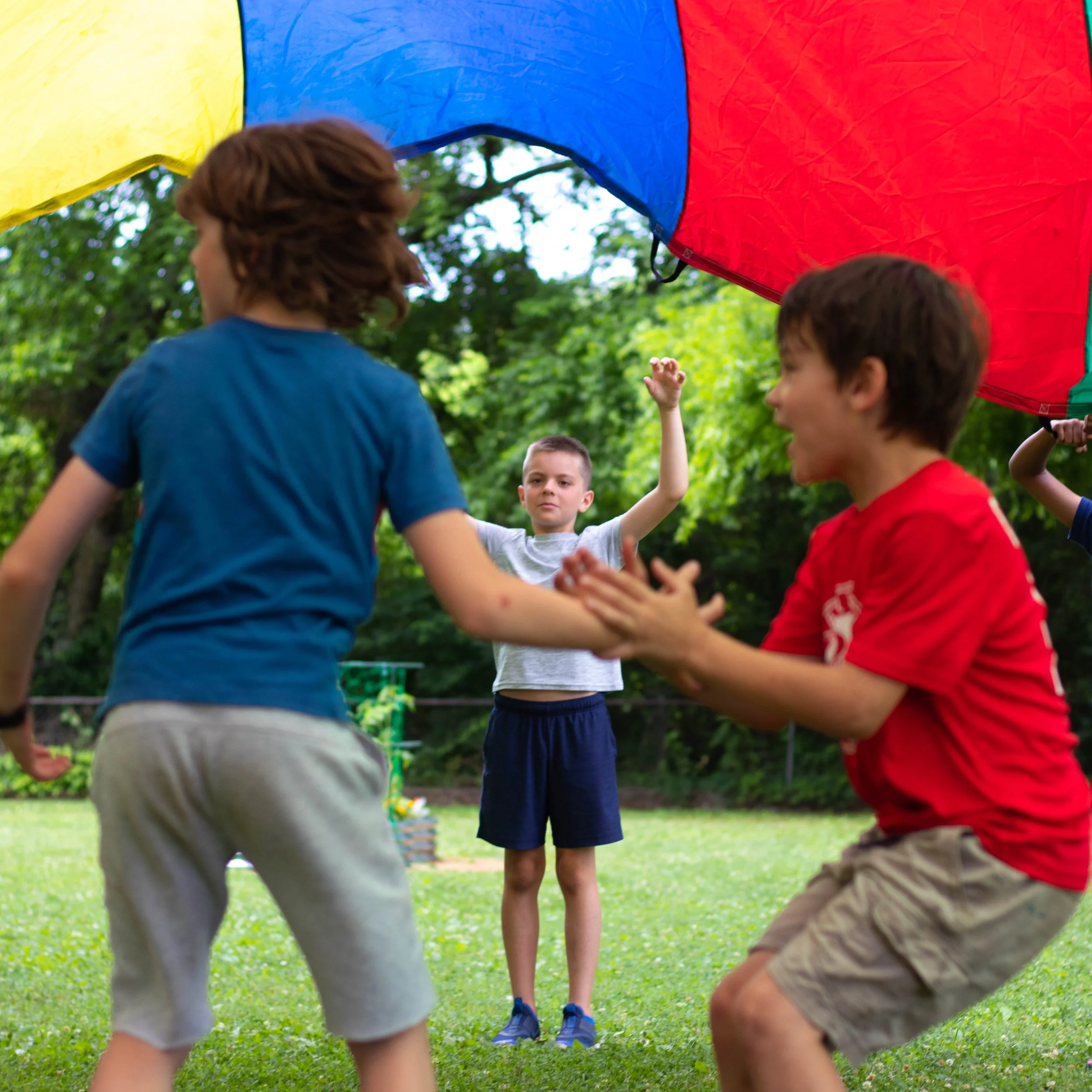 Children playing under a colorful parachute outdoors on a grassy field with trees in the background.