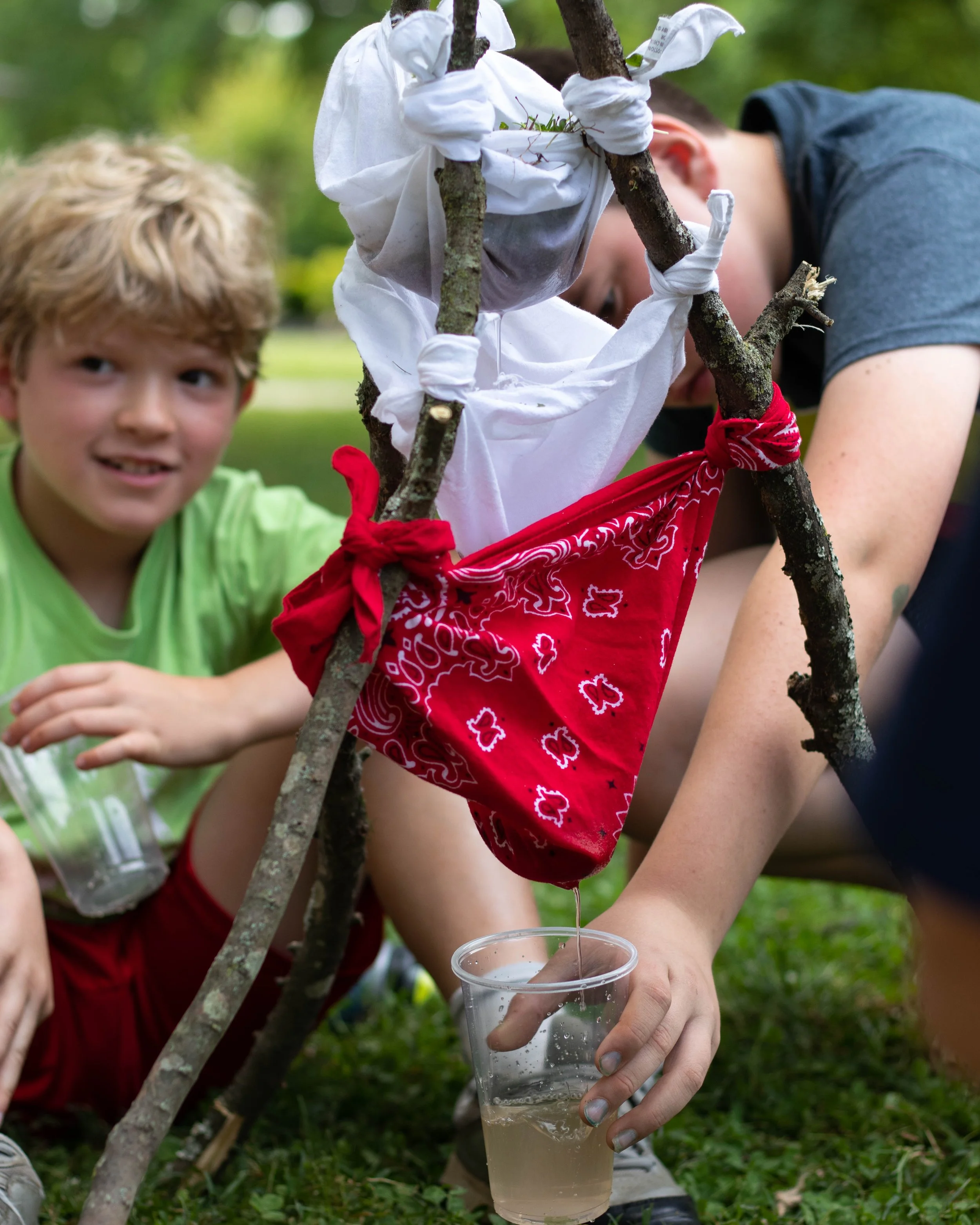 Two children collect rainwater in a plastic cup from a rain-catching device made of sticks and fabric, outdoors in a park.