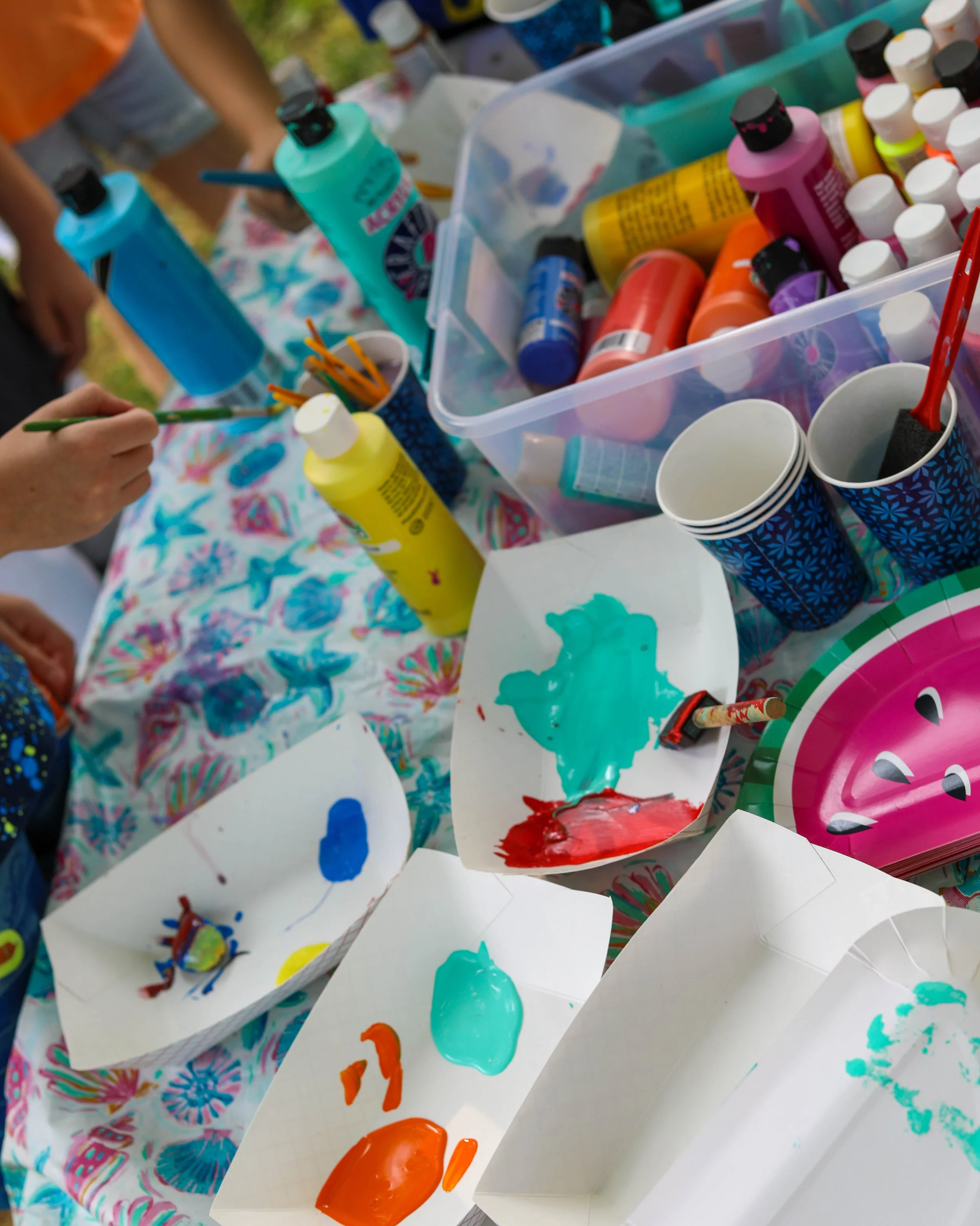 A table covered with a colorful tablecloth and various art supplies, including bottles of paint in different colors, paper cups, paintbrushes, and paper plates with mixed paint. Multiple hands are seen holding brushes, engaging in finger painting or mixing paint.