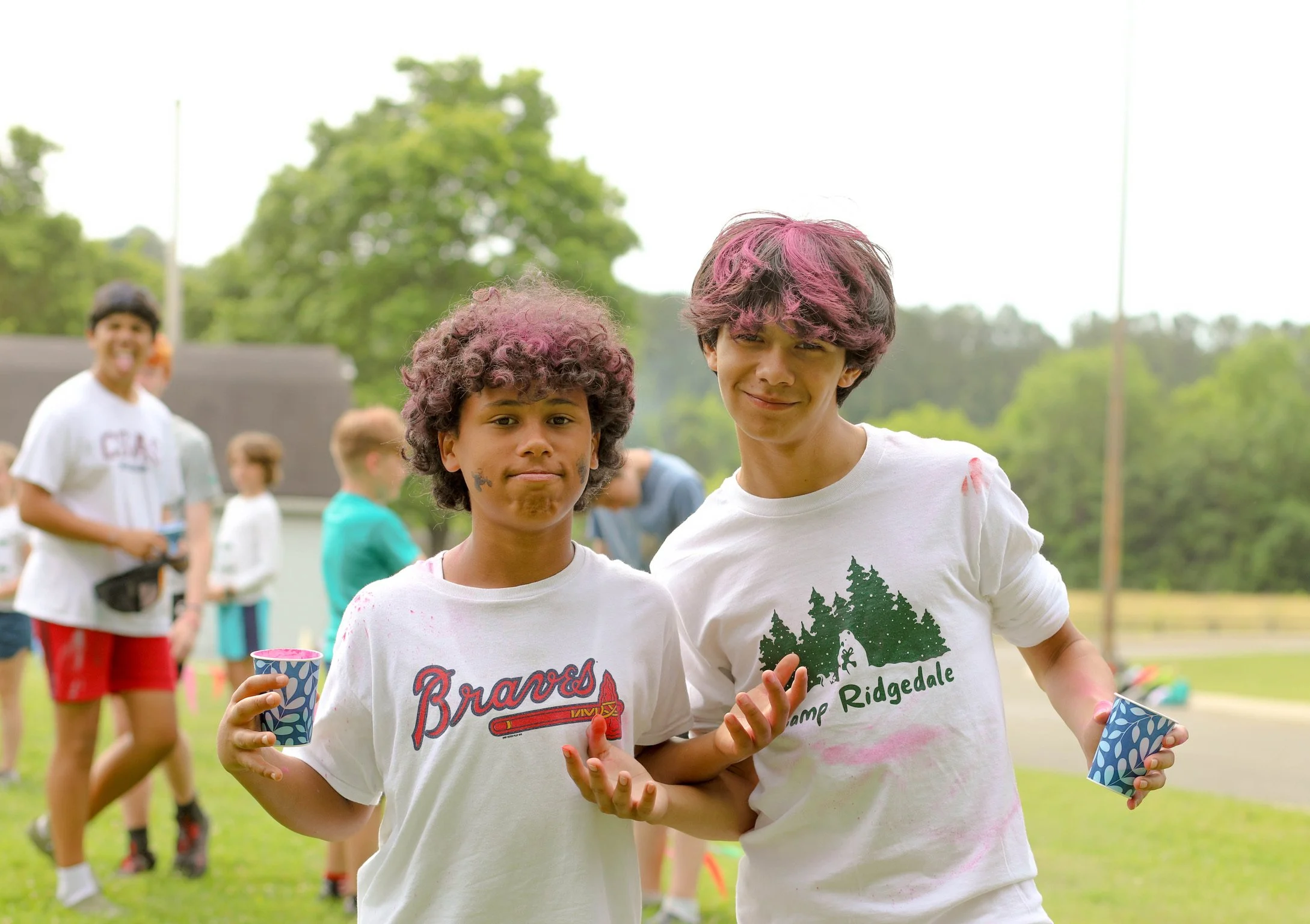 Two boys with colored hair and paint on their faces, holding cups, standing outdoors with other kids in the background at a camp or outdoor event.