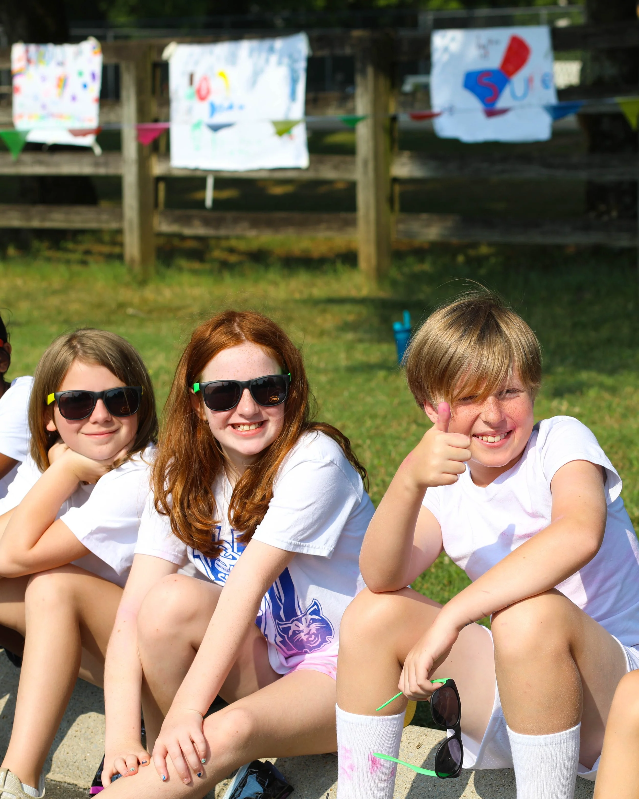 Three kids sitting outside on a sunny day, with colorful banners hanging on a wooden fence in the background. They are smiling and posing for the photo, wearing white T-shirts and sunglasses.