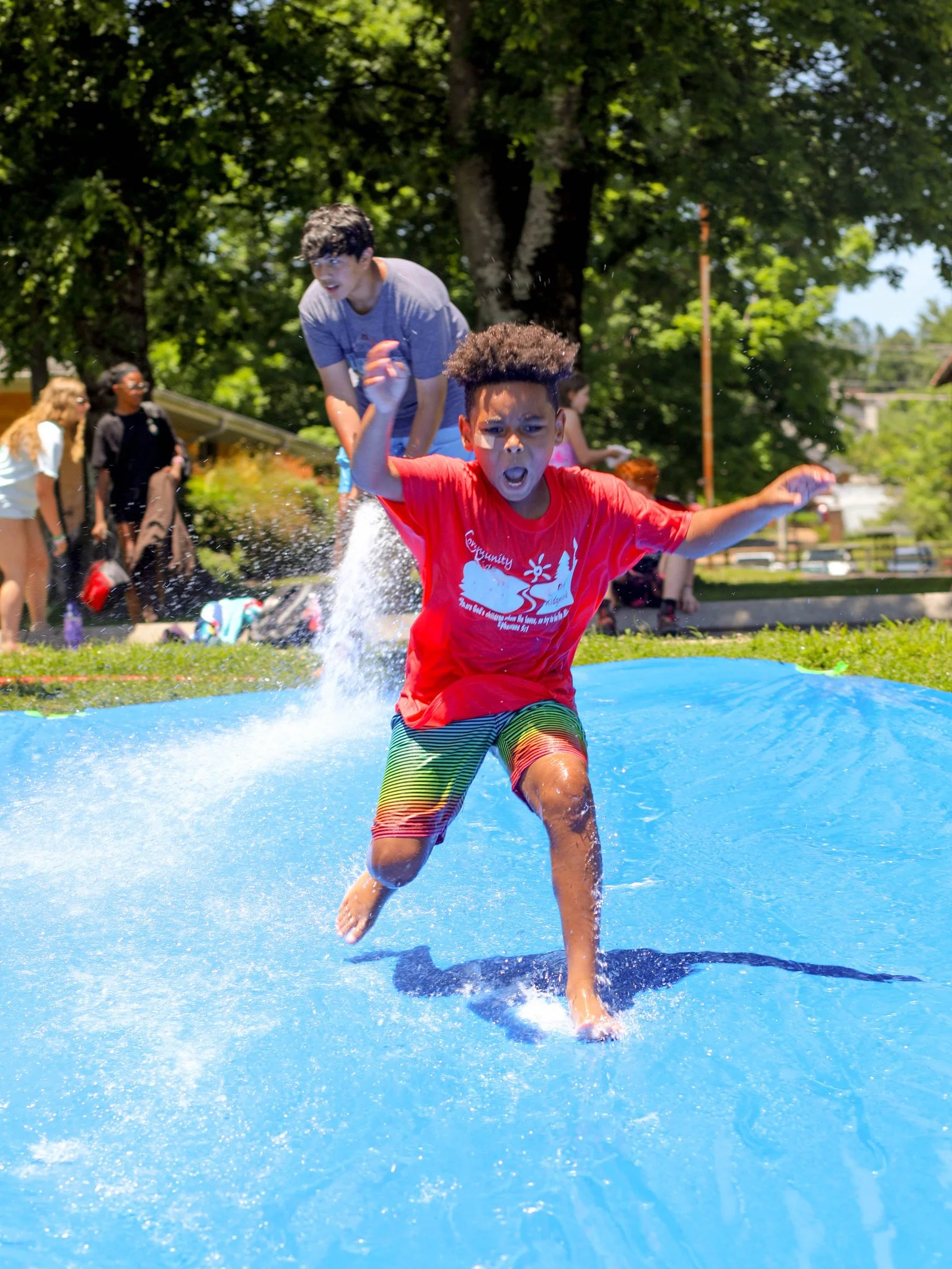 Child wearing red shirt and striped shorts running on water in a splash pad, with people and trees in the background.