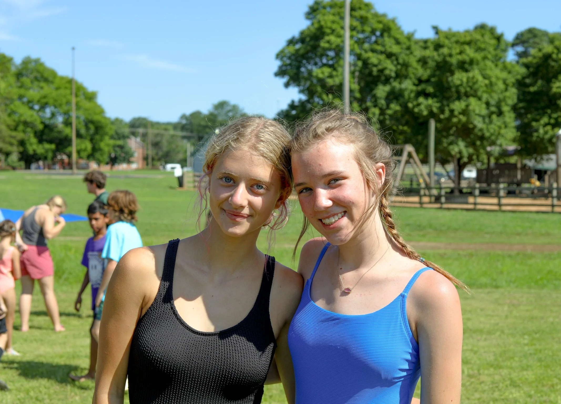 Two young women smiling outdoors on a sunny day with green trees and a playground in the background.