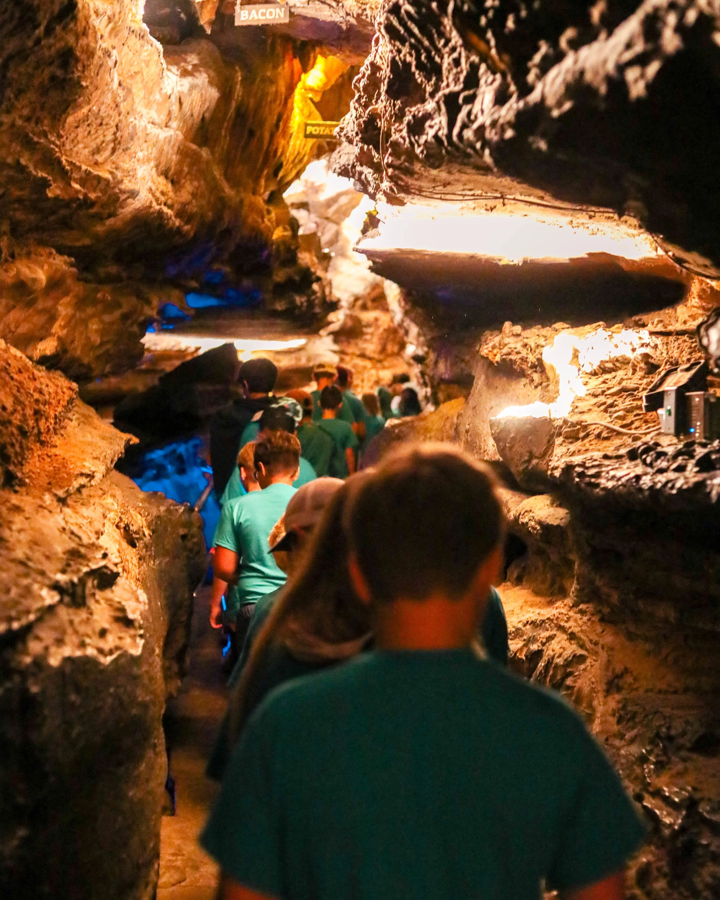 A group of people walking through an underground cave with rocky walls and dim lighting, some signs indicating sections such as 'BACON' and 'POTATO'.