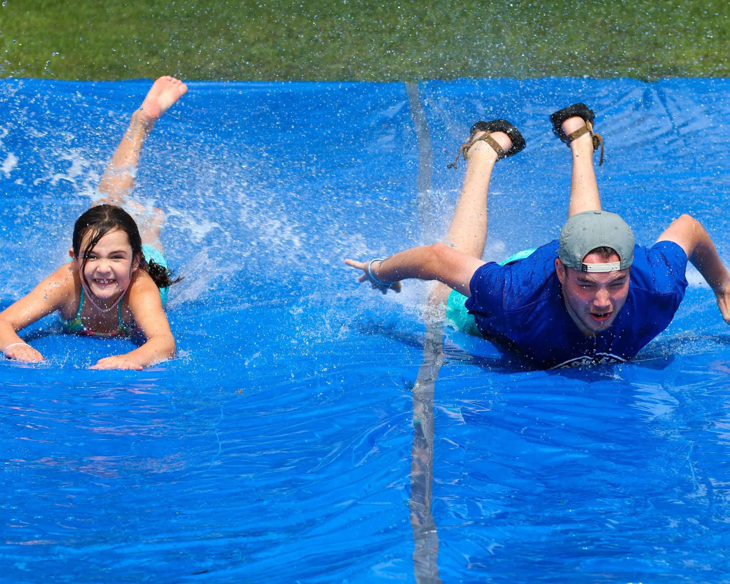 A young girl and a man slide down a water slip-and-slide on a sunny day, creating splashes of water and smiling.