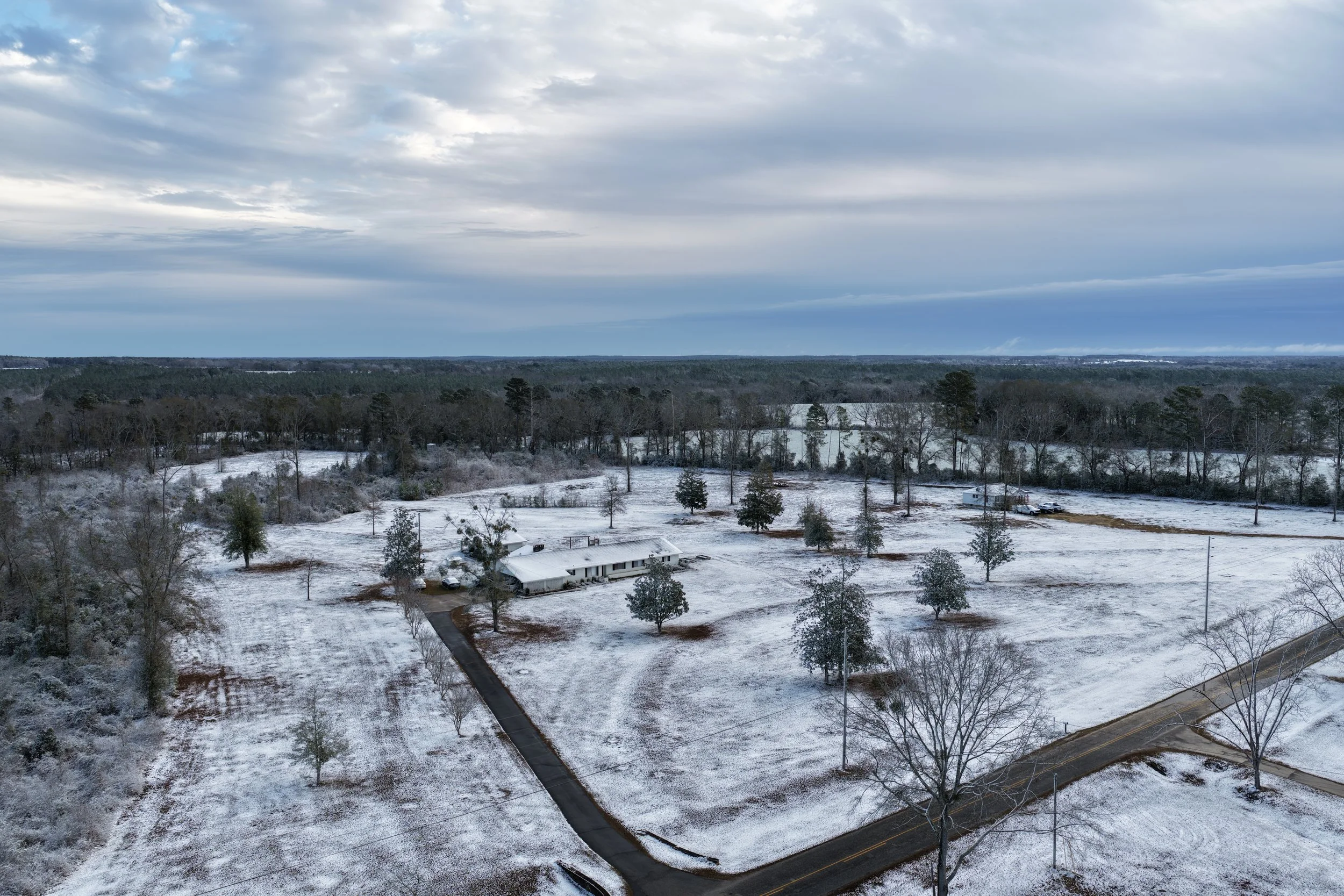 A snow-covered rural landscape with a house, trees, and a winding road under a cloudy sky.