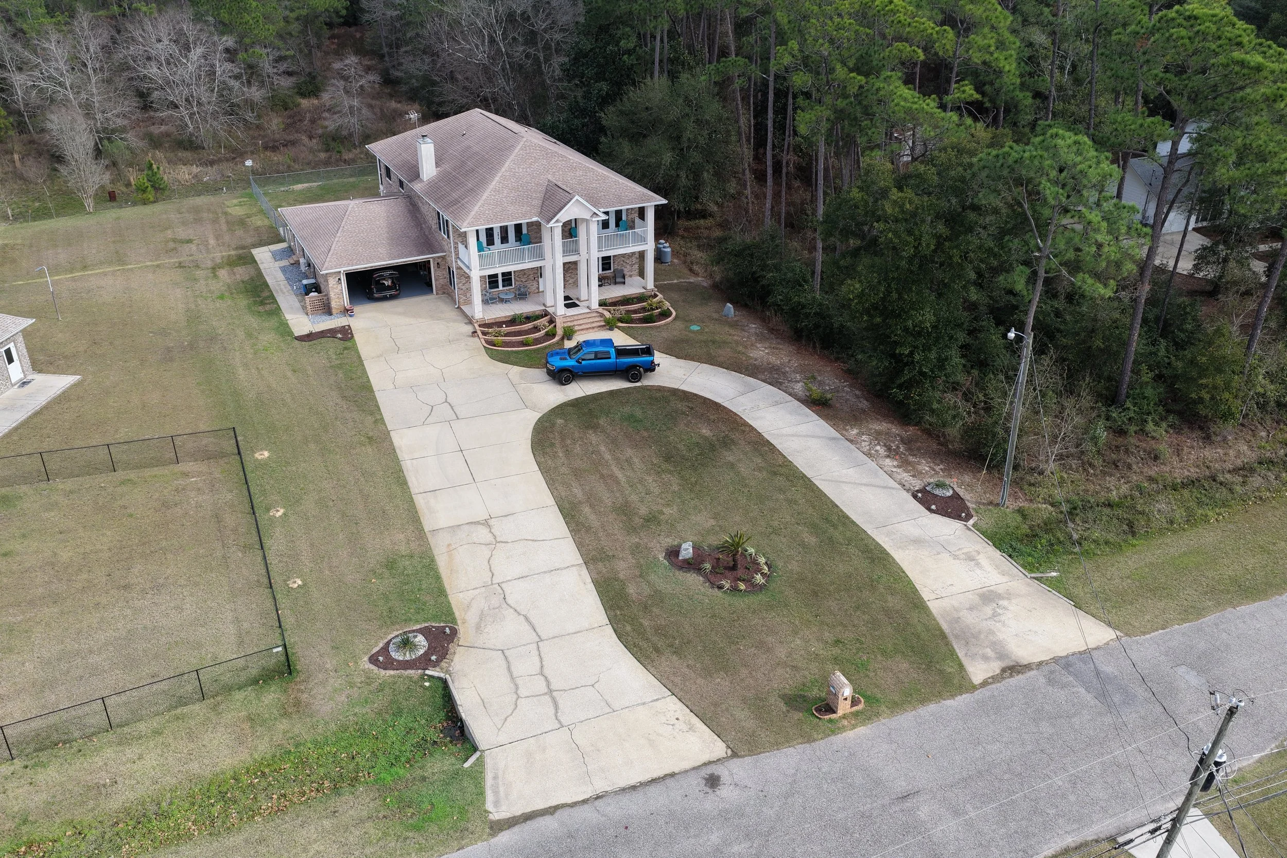 An aerial view of a two-story house with a front porch, a garage with a black car, and a bright blue truck parked on the driveway. The house has a landscaped yard with small flower beds, and is surrounded by a grassy lawn and a wooded area with tall trees in the background.