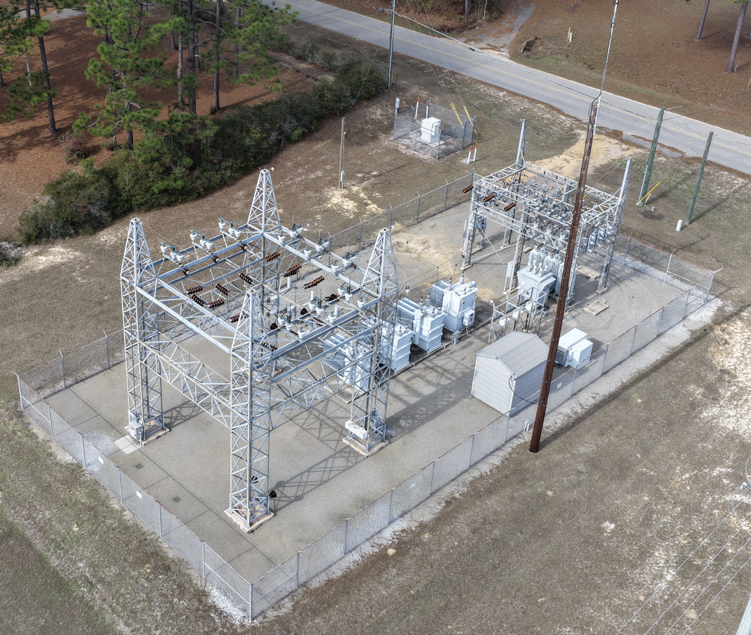 Aerial view of an electrical substation with metal framework, transformers, insulators, and surrounding fencing, situated near a road with trees and grass nearby.