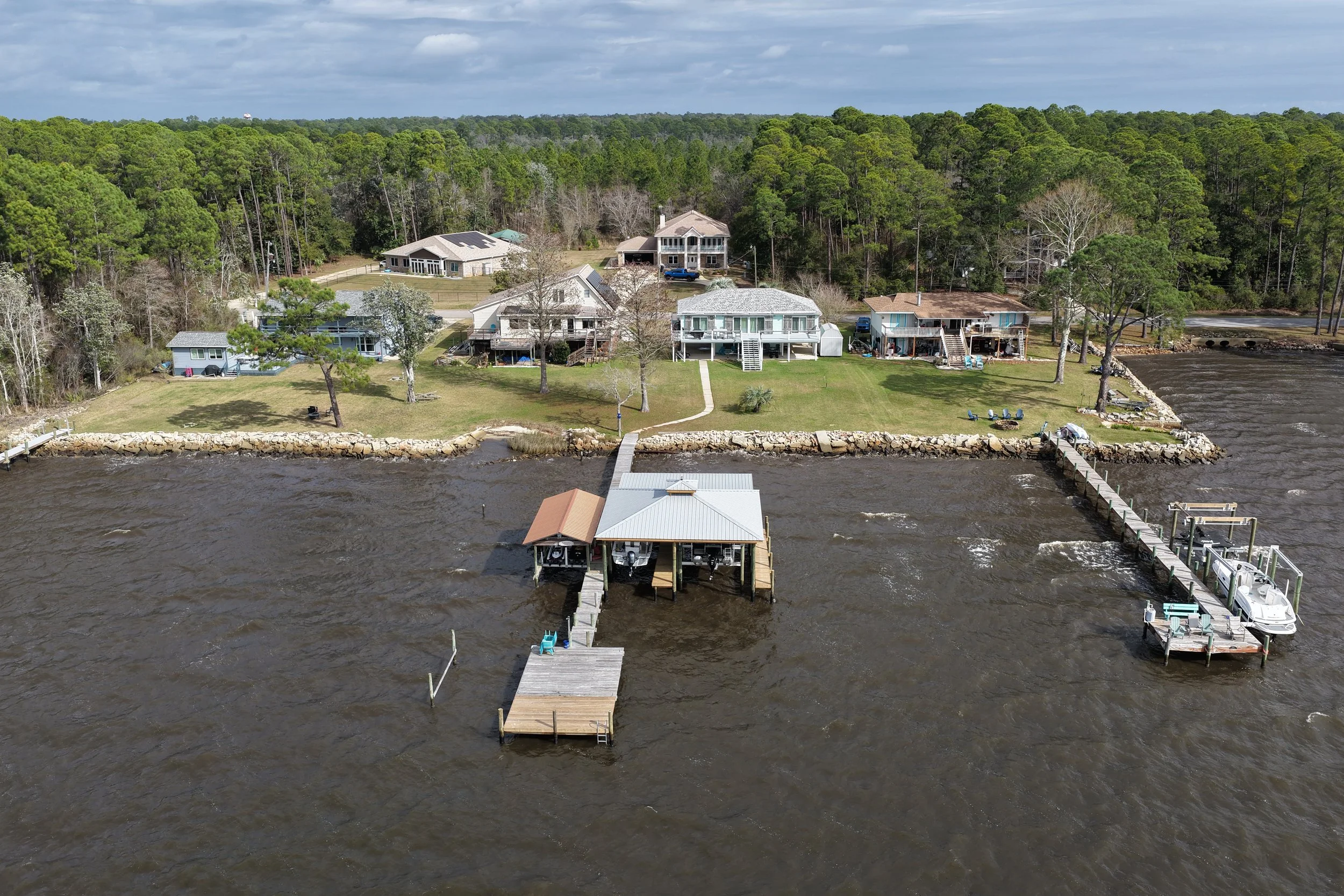Aerial view of waterfront houses with boat docks on a river, green trees behind the houses, with a cloudy sky.