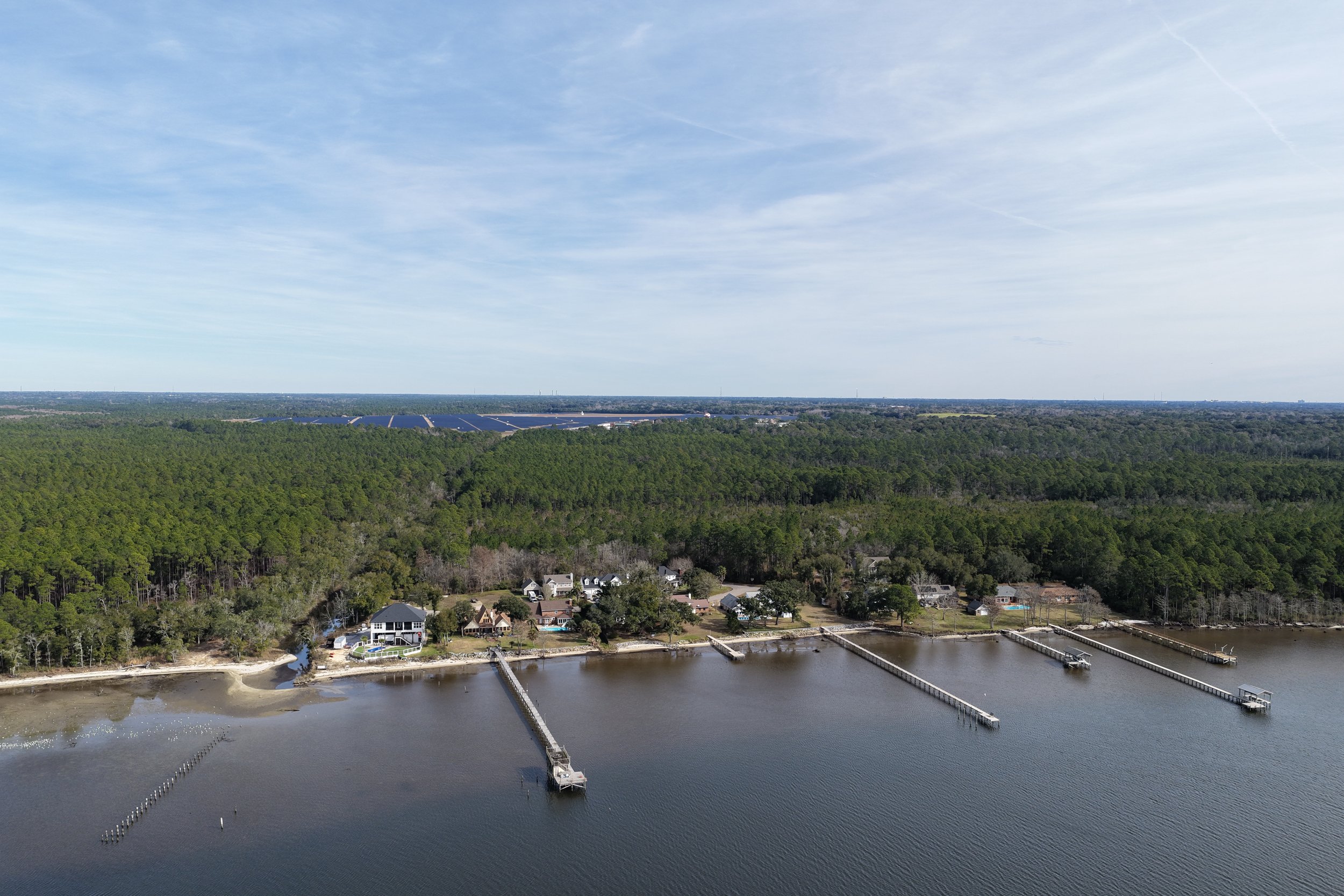 Aerial view of a waterfront community with houses along the shoreline, multiple docks extending into the water, and a large wooded forest in the background under a partly cloudy sky.