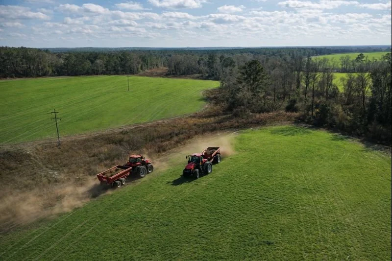 Two red tractors are plowing a grassy field, kicking up dust in a rural landscape with green fields, trees, and a partly cloudy sky.