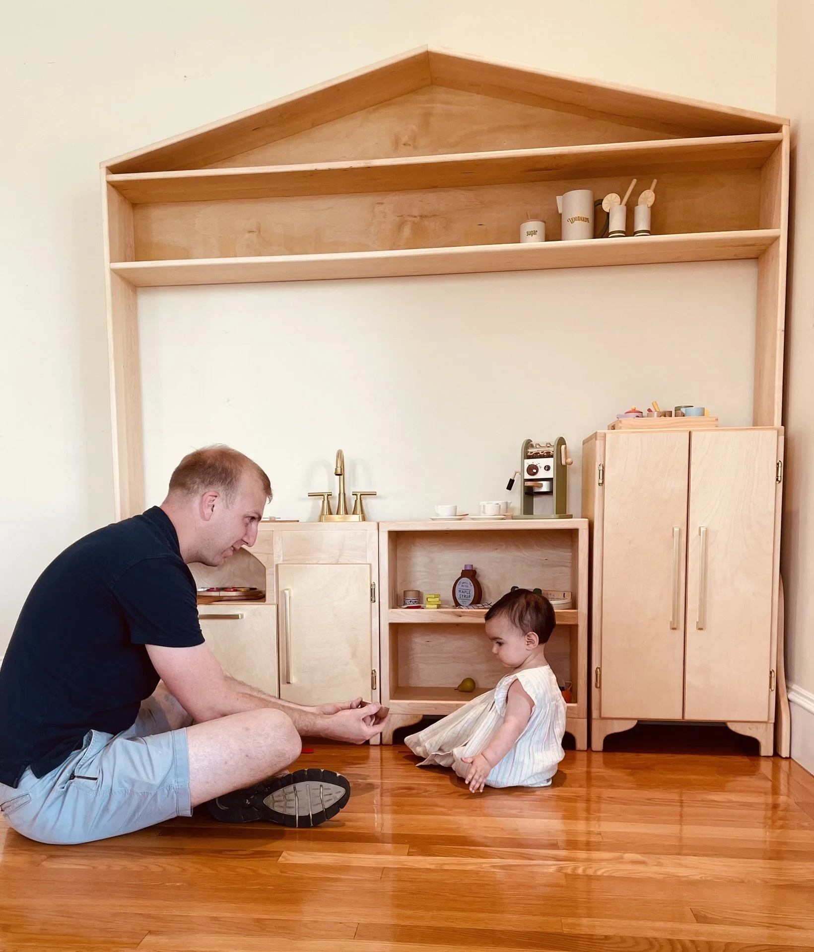 A man and a young girl sitting on a hardwood floor in front of a custom wooden play Kitchen, playing together. The room has a beige wall and a large wooden decorative shelf above the play area.