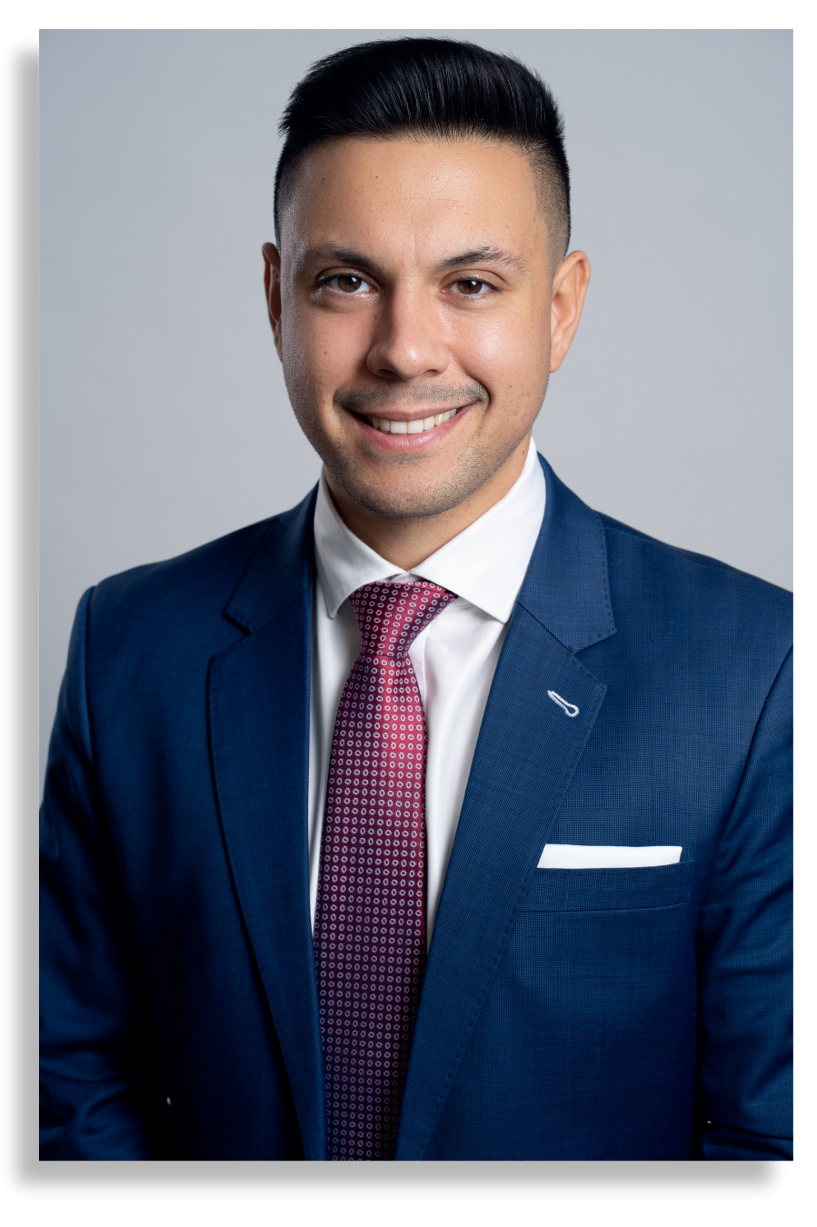 Professional headshot of a man in a blue suit, white shirt, and red patterned tie, smiling against a gray background.