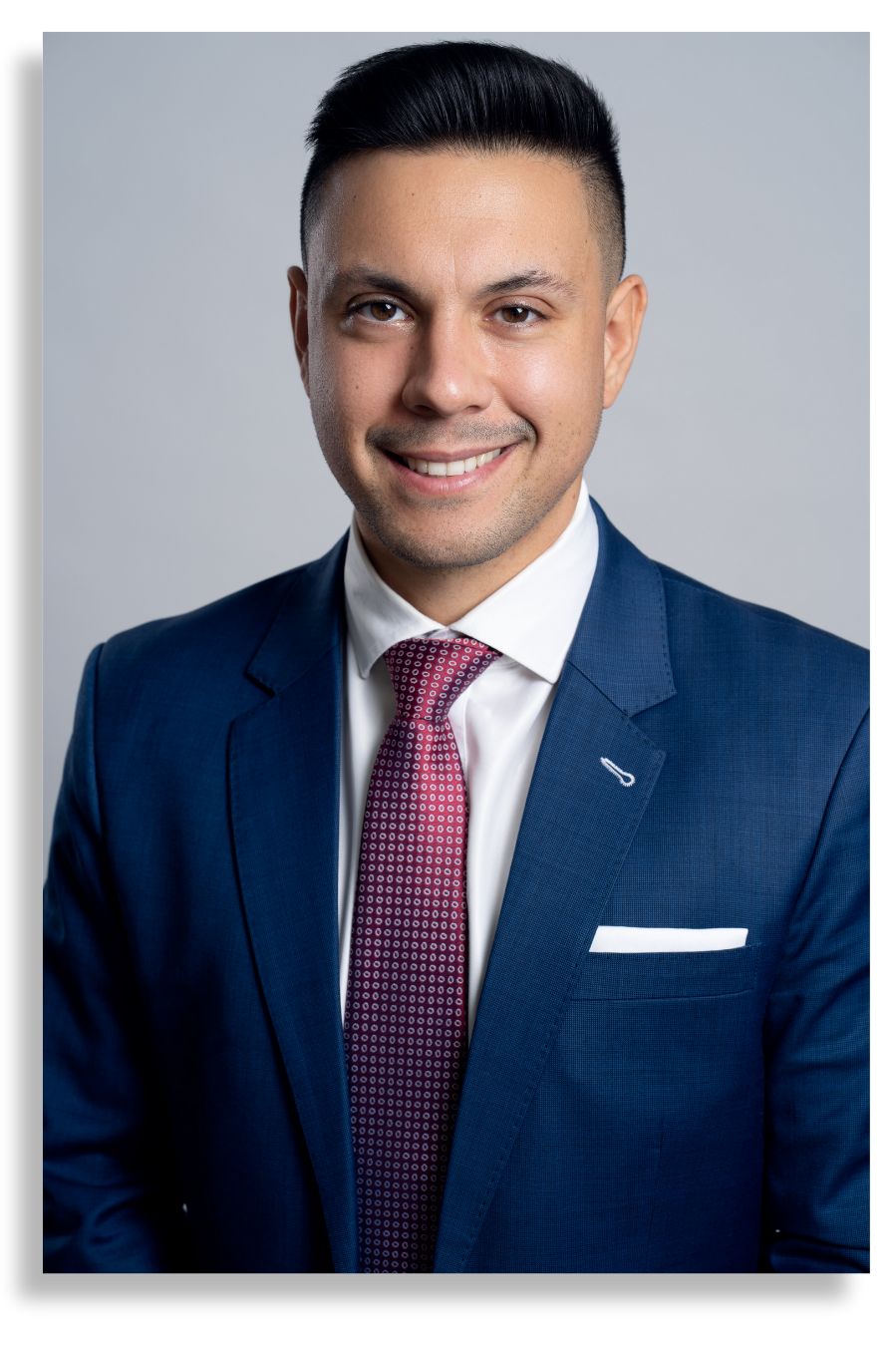 A young man in a blue suit with a pink patterned tie and white shirt, smiling against a gray background.