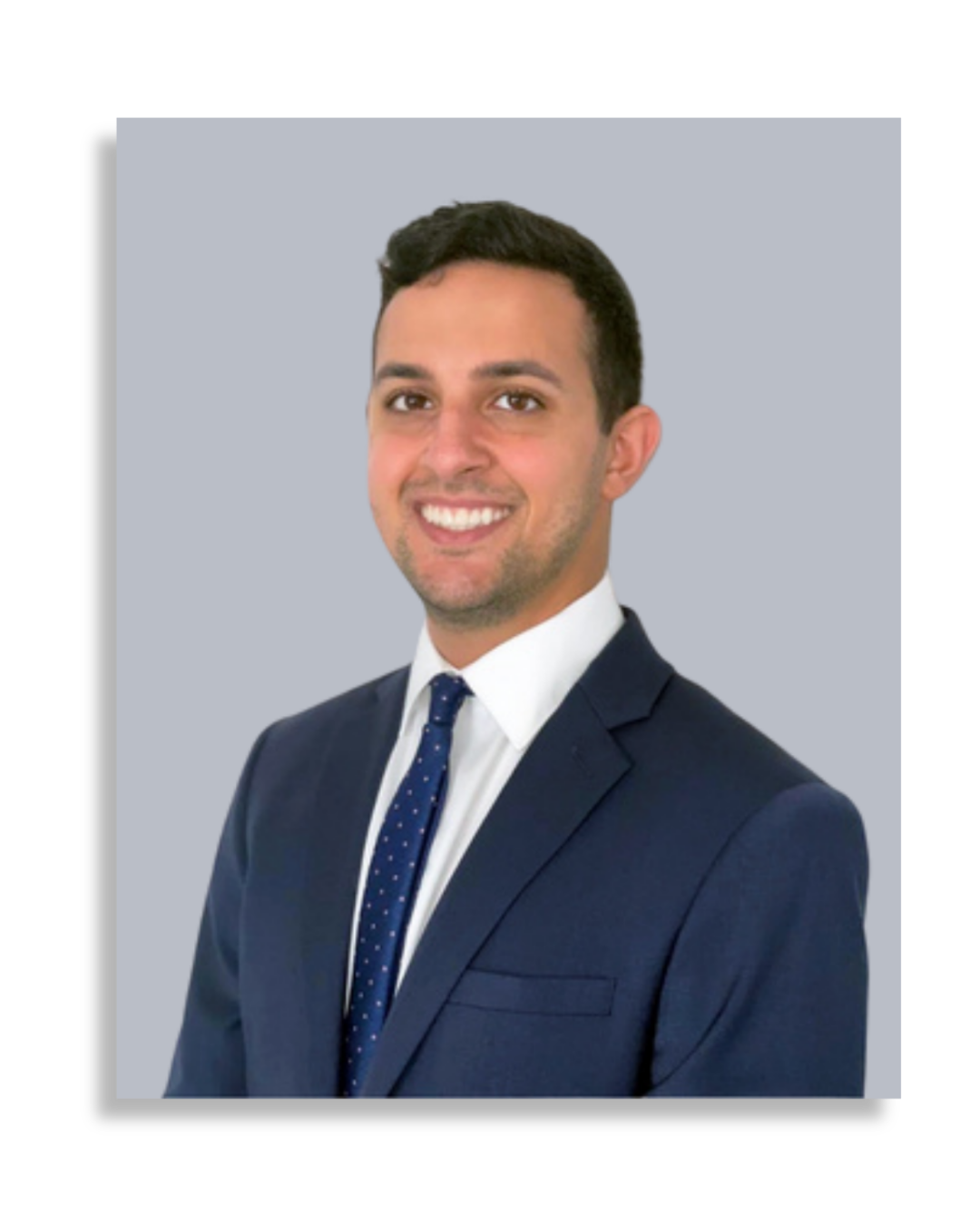 Professional headshot of a smiling man in a navy suit, white shirt, and navy tie with white polka dots, against a light gray background.