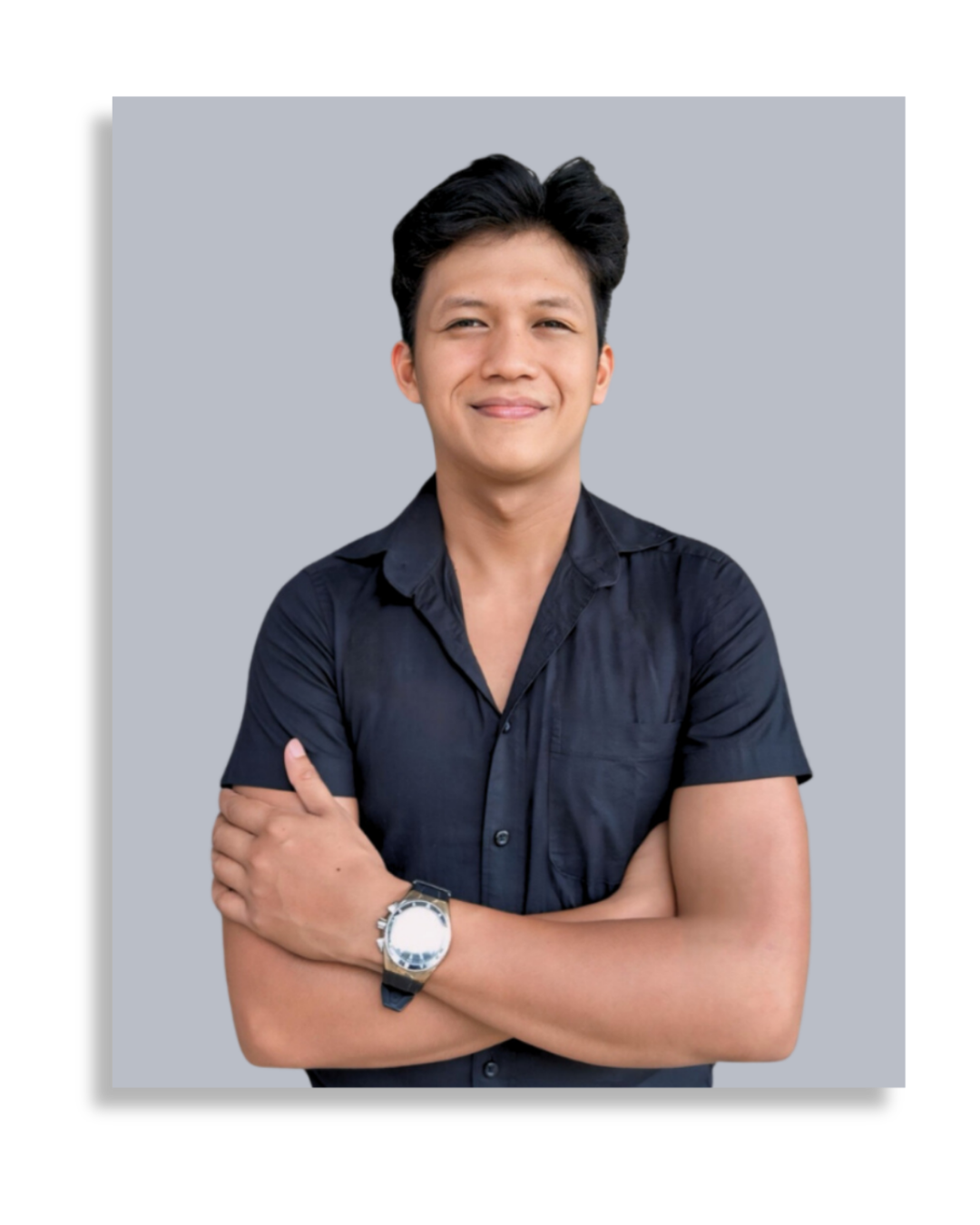 Young man with black hair, smiling, wearing a navy blue shirt, crossing arms, silver watch on left wrist, against a light gray and black background.