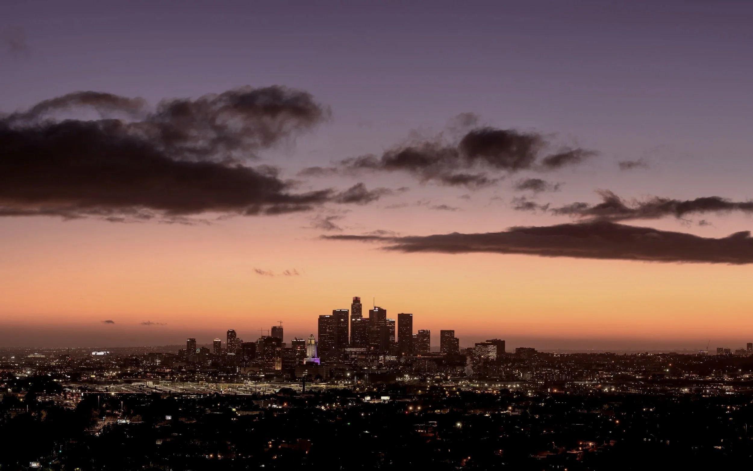 City skyline at sunset with dark clouds overhead and city lights beginning to twinkle.
