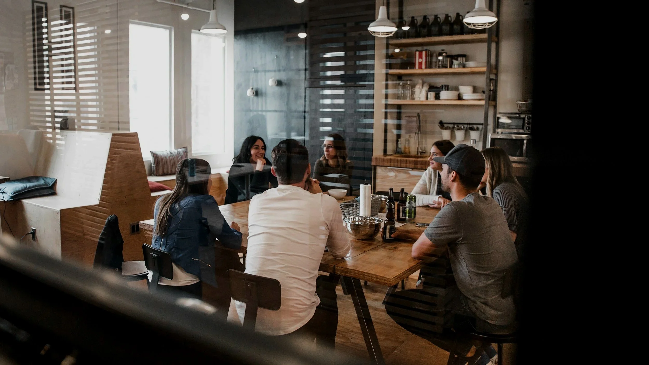 Group of seven friends sitting around a wooden table in a cozy, modern restaurant, engaged in conversation and smiling. The restaurant features open shelving with plates and glasses on the wall, large windows with blinds, and ambient lighting.