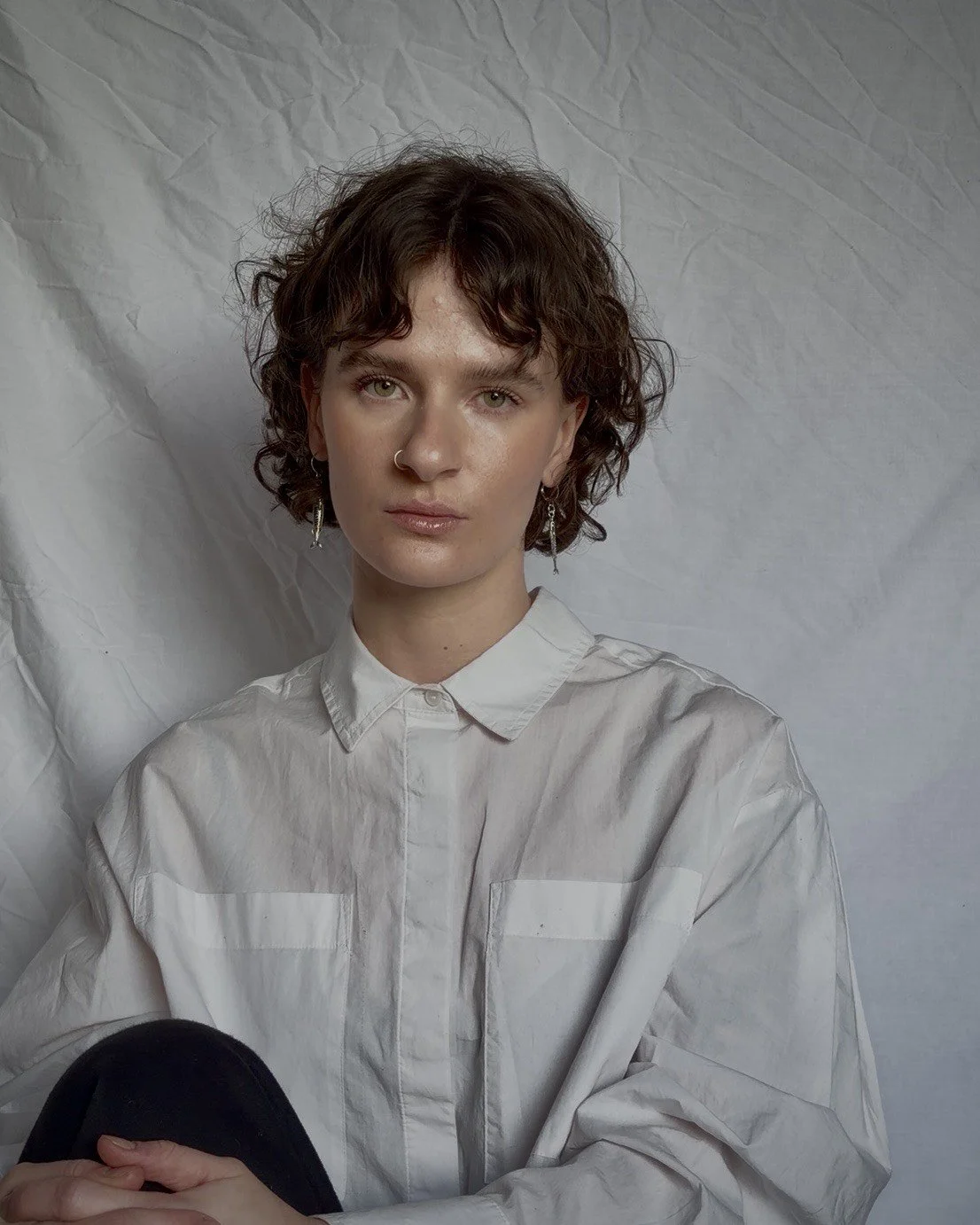 Portrait of a woman with short, curly brown hair, wearing a white button-up shirt, earrings, and a nose ring, sitting against a crumpled white background.