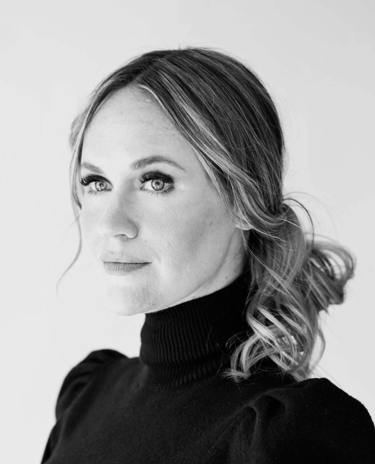 Black and white portrait of a woman with wavy hair wearing a black turtleneck sweater, looking to the side.