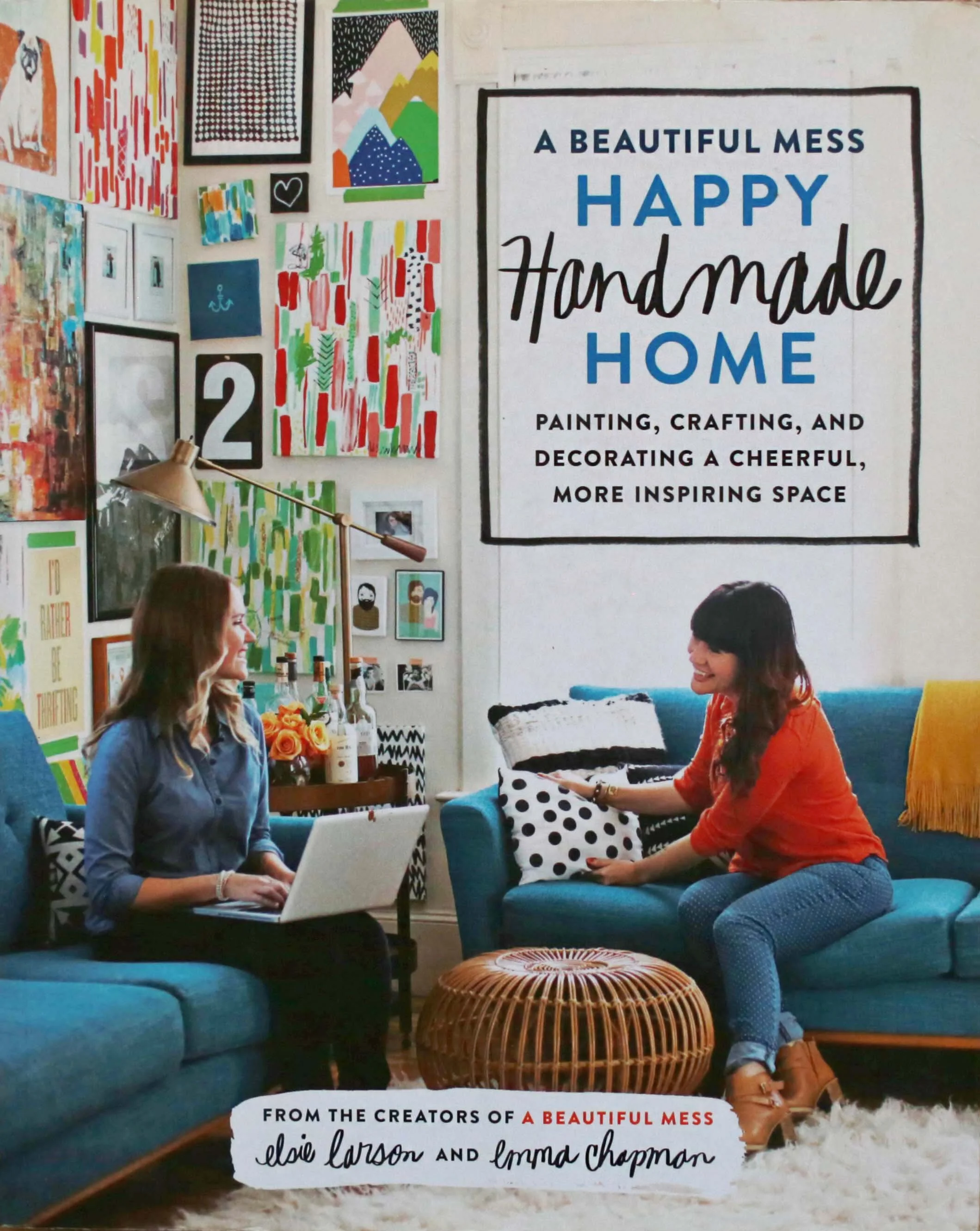 Two women sitting and smiling in a colorful, decorated living room with art on the walls, a large sign reading 'A Beautiful Mess, Happy Handmade Home,' and a wicker coffee table between them.