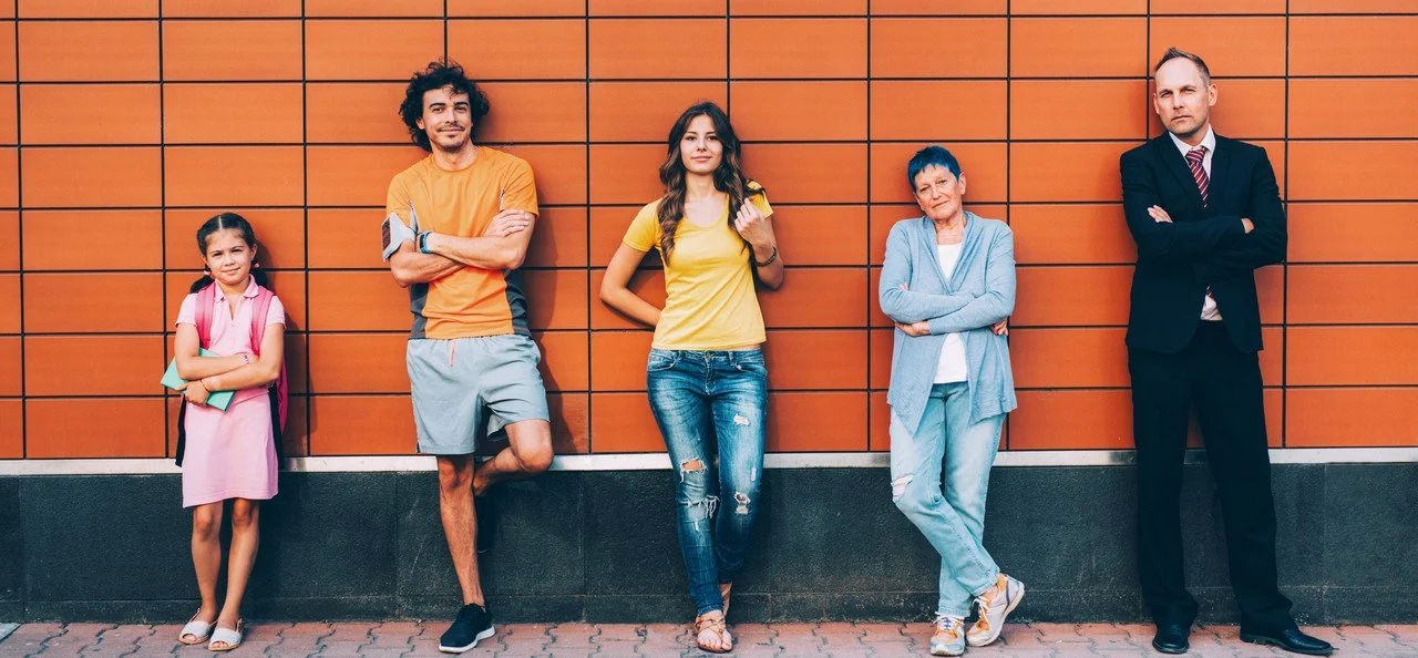 Fünf Menschen stehen an einer orangefarbenen Wand, voneinander entfernt, im Freien.
