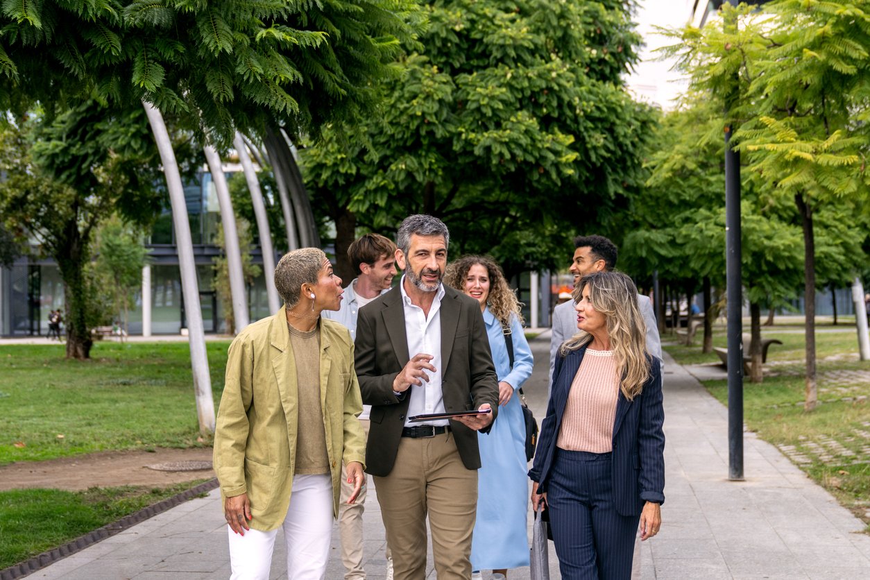 Eine Gruppe von sieben berufstätigen Menschen, die sich in einem Park in der Stadt unterhalten, bei gutem Wetter.