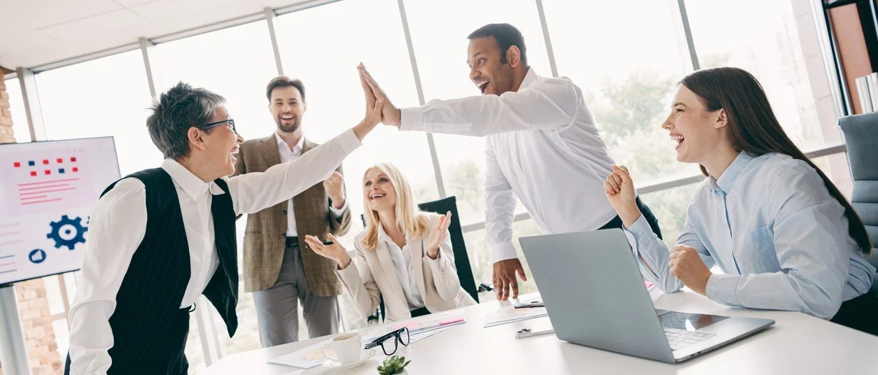 Geschäftsleute feiern einen Erfolg im Büro durch High-Five und Lachen, mit Laptop, Dokumenten und einem Präsentationsbildschirm im Hintergrund.