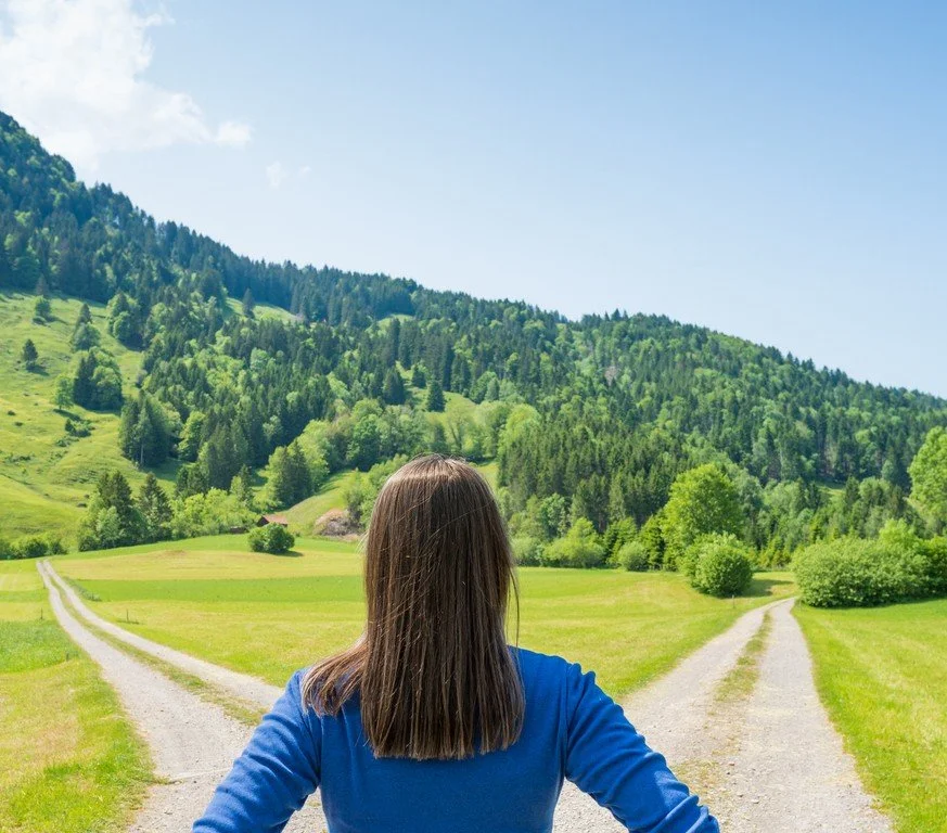 Eine Frau steht vor zwei Weggabelungen in einer grünen Wiese mit Bäumen und Hügeln im Hintergrund, bei sonnigem Himmel.