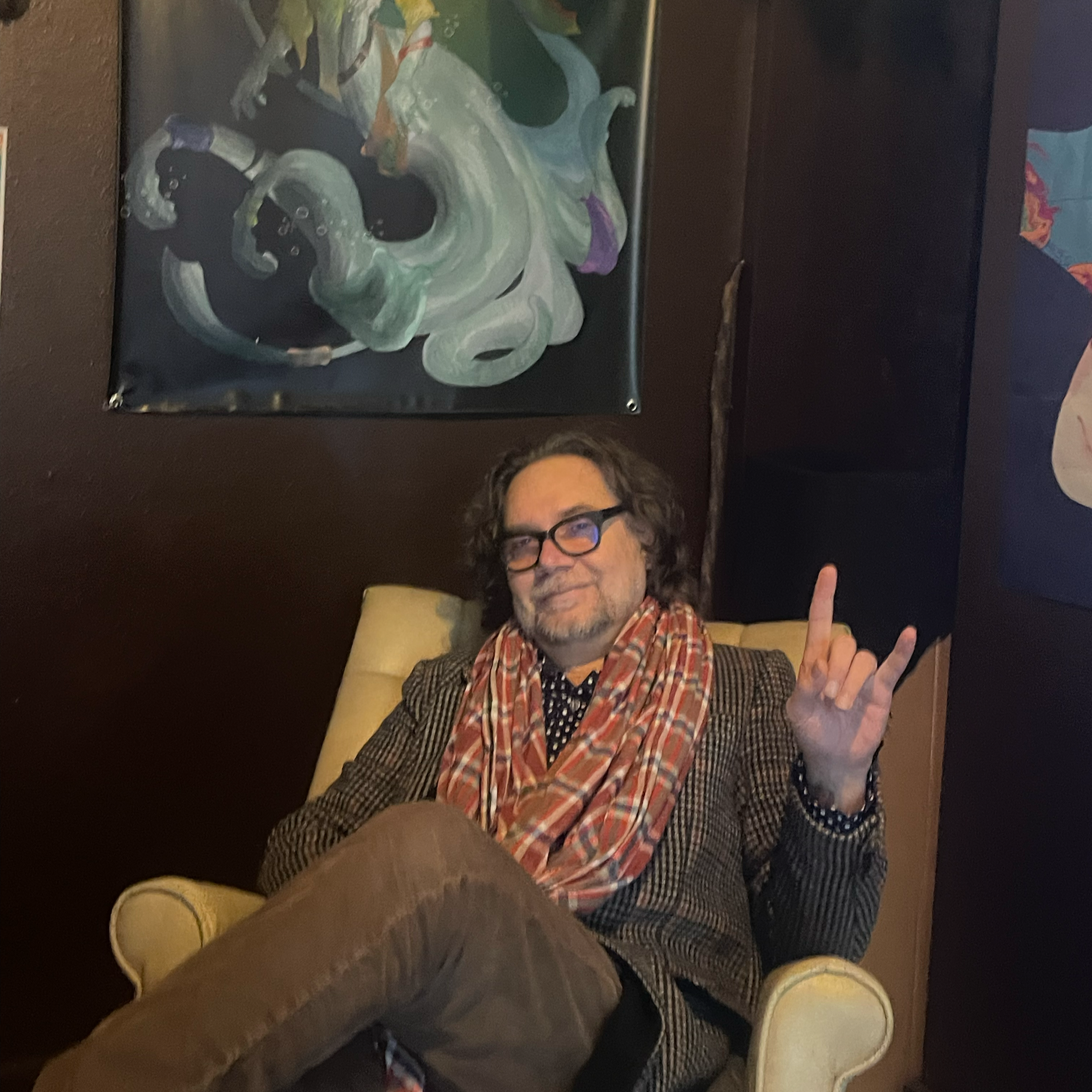 A man with curly hair, glasses, and a beard sitting in a yellow armchair, wearing a patterned blazer and a scarf, making a rock and roll hand sign, with abstract paintings hanging on dark-colored walls behind him.