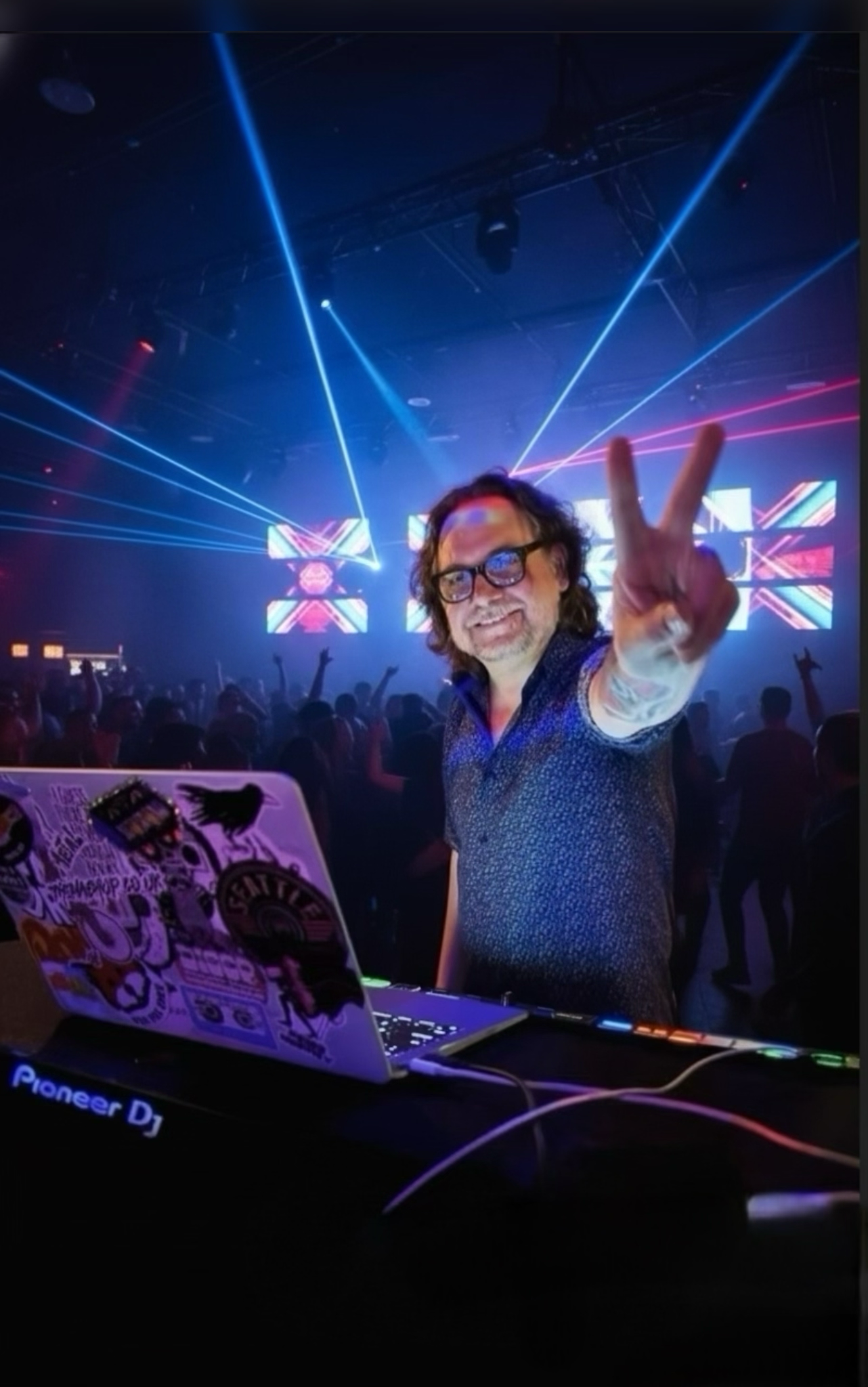 A man with glasses smiling and making a peace sign in front of a DJ setup at a nightclub with colorful lights and a crowd in the background.