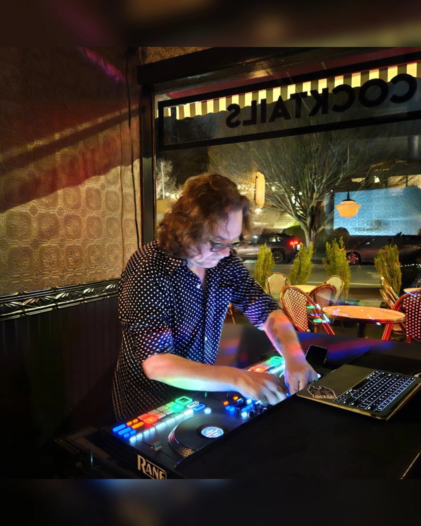 A DJ in a polka dot shirt using a digital DJ controller at a bar or restaurant with colorful LED lights, a laptop, and a window showing cars and trees outside at night.
