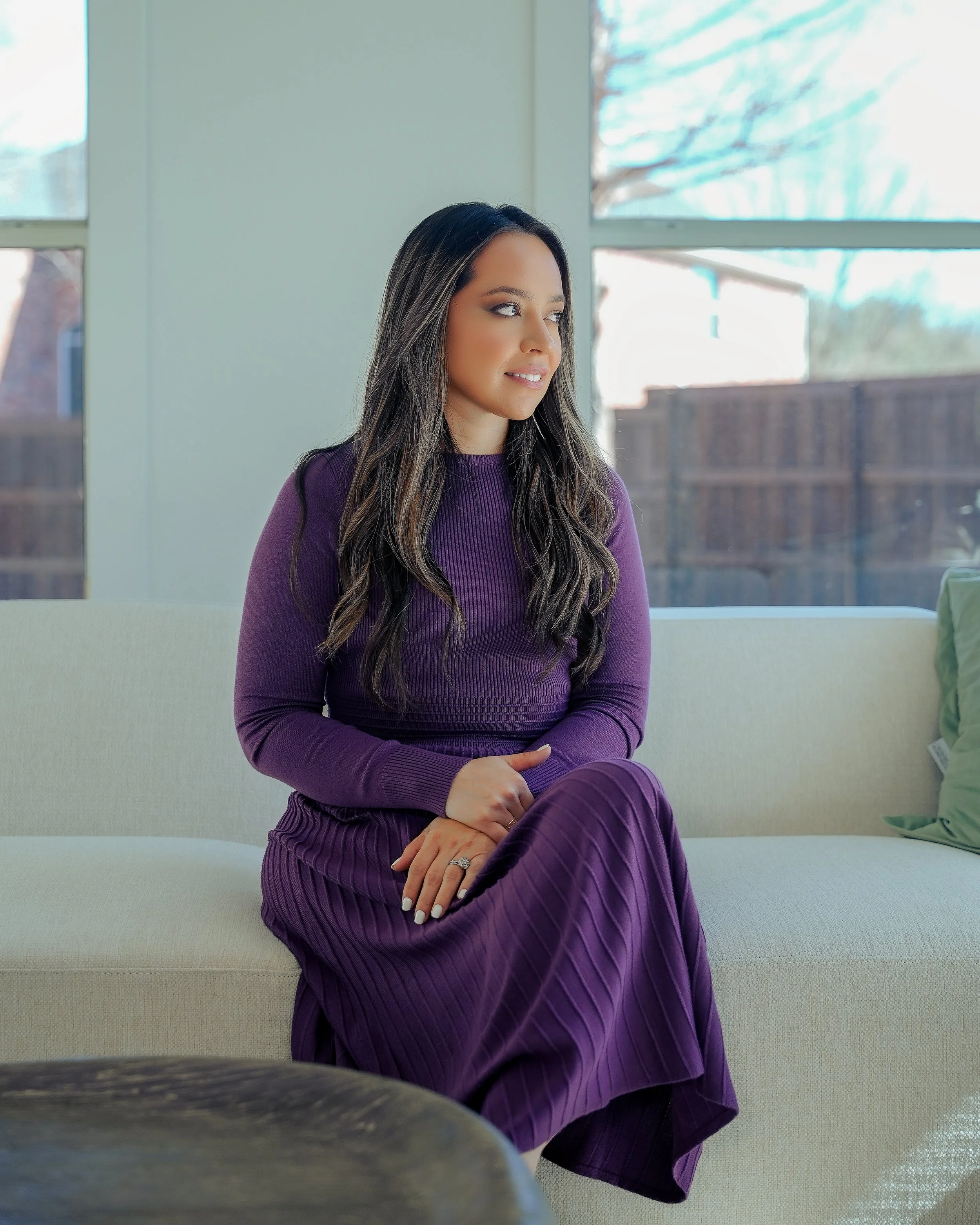 A woman with long dark hair sitting on a cream-colored sofa, wearing a purple dress, in a room with large windows showing a fenced backyard and trees outside.