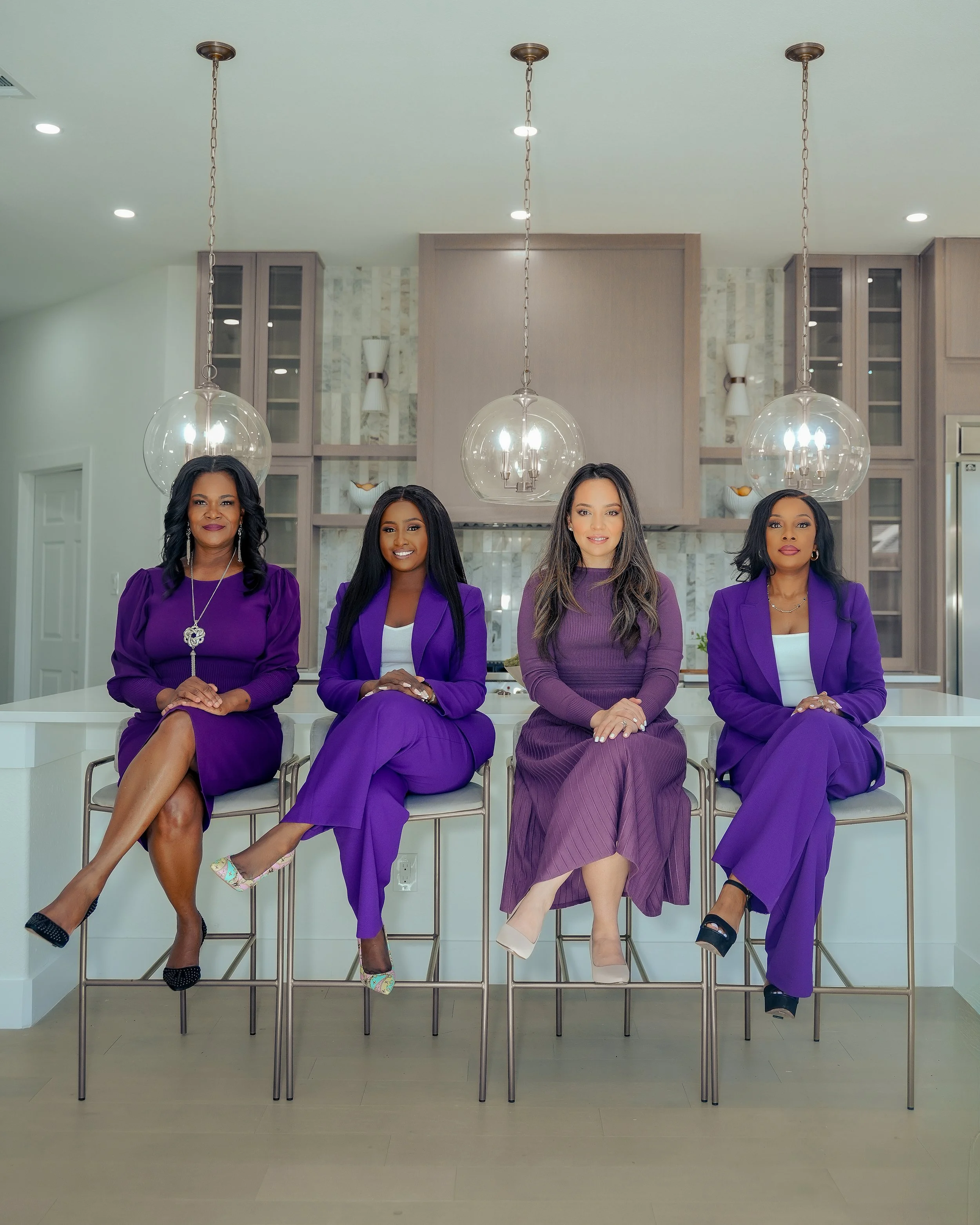 Four women dressed in purple suits and dresses sitting on bar stools in a modern kitchen.