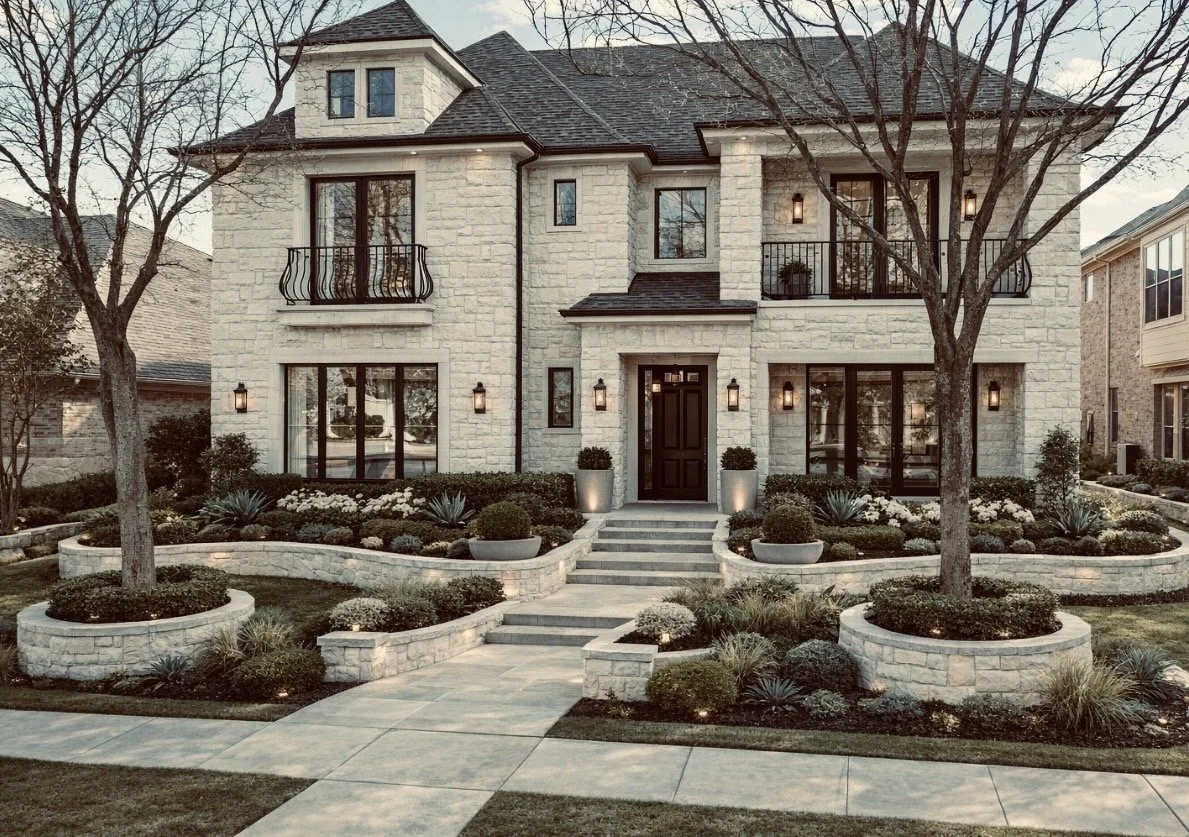 Front view of a luxurious two-story house with white stone exterior, black roof, and black window frames. The house features two balconies with black railings, multiple large windows, and a black front door. The landscaped yard has trimmed bushes, small trees, potted plants, and a paved walkway with stairs leading to the front entrance. The property is illuminated with outdoor wall-mounted lights.