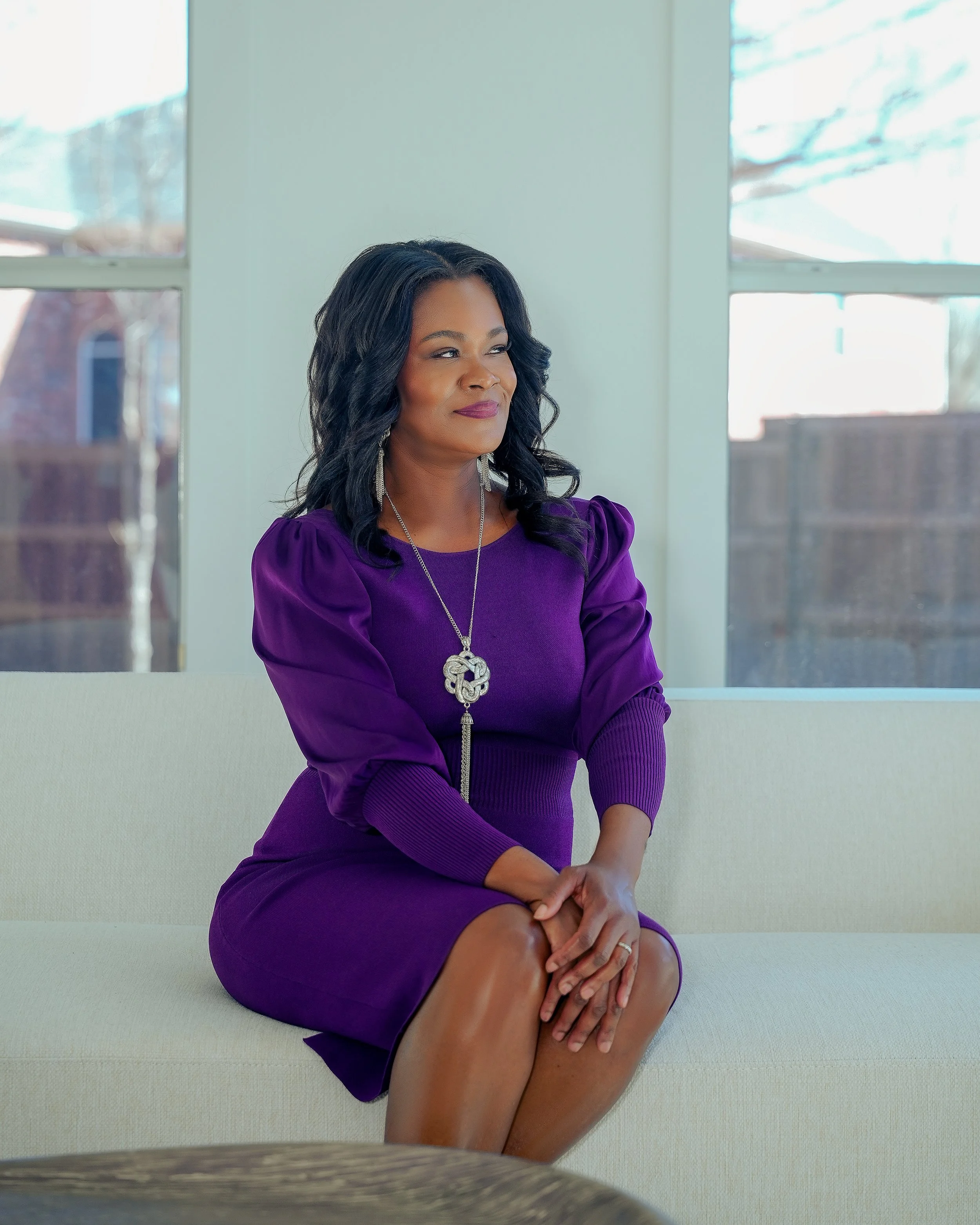 A woman with dark, wavy hair and a dark complexion sitting on a beige sofa indoors, wearing a purple dress with puffy sleeves and a large pendant necklace, with windows in the background showing trees and a building.