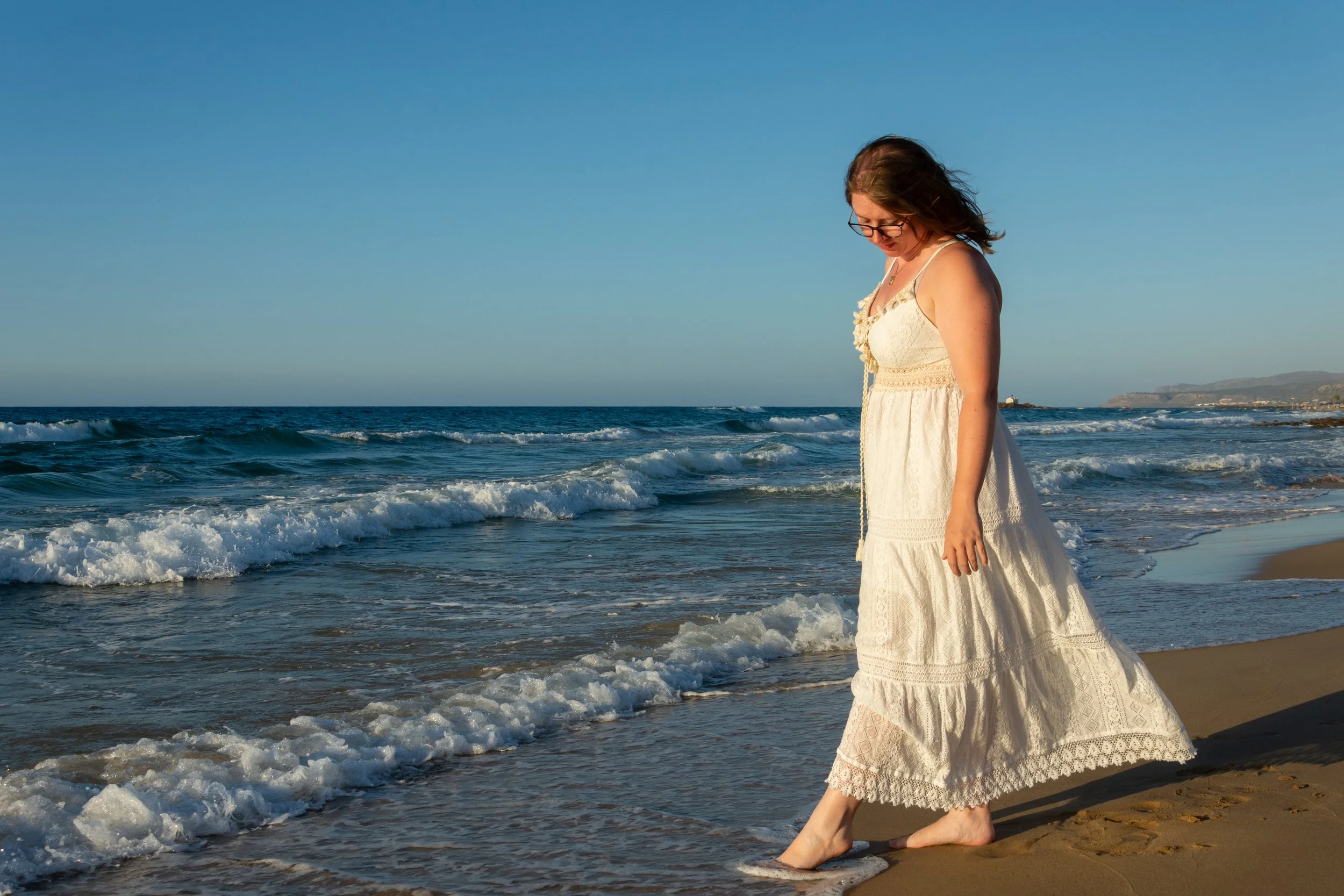 Une femme en robe blanche se tient pieds nus sur la plage, regardant l'eau de l'océan au coucher du soleil.