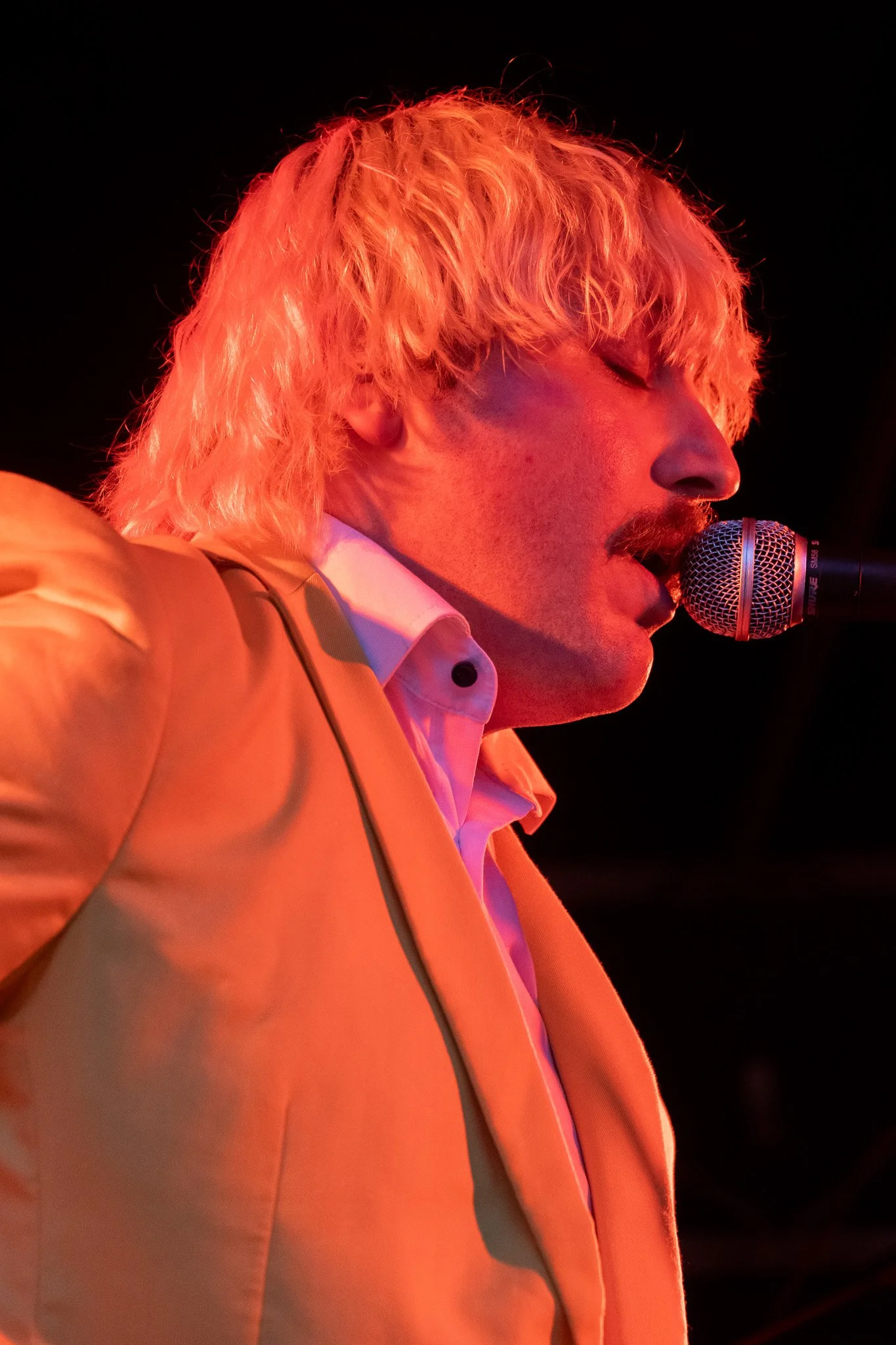 Un homme avec des cheveux blonds et ondulés, portant un costume beige clair et une chemise blanche, chante ou parle dans un microphone, en éclairage rouge-orange dans un environnement sombre.