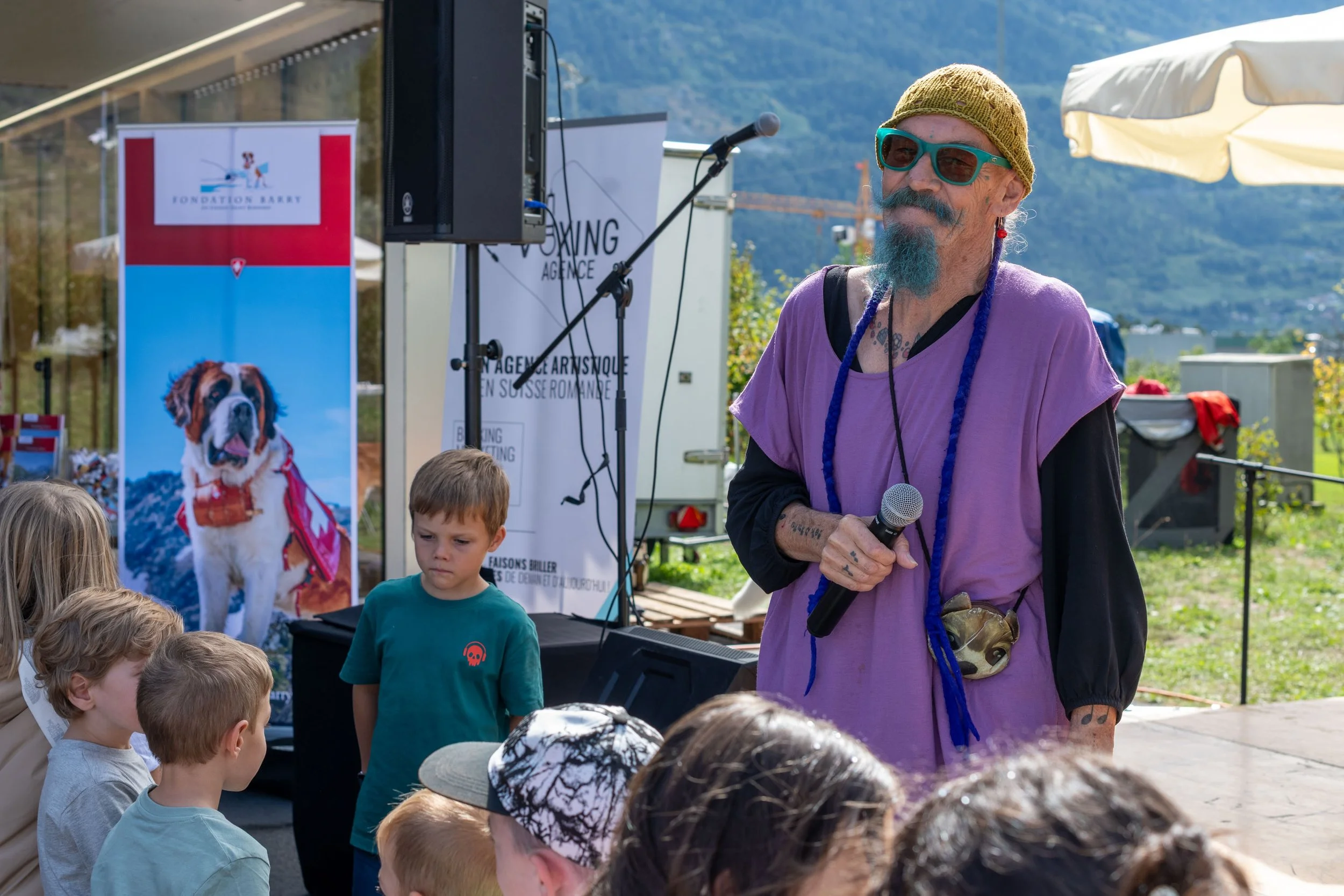 Un homme avec une barbe bleue, portant des lunettes de soleil, un bonnet beige, et une tenue violette, tient un microphone lors d'un événement extérieur, avec un groupe d'enfants devant lui et des affiches en arrière-plan.