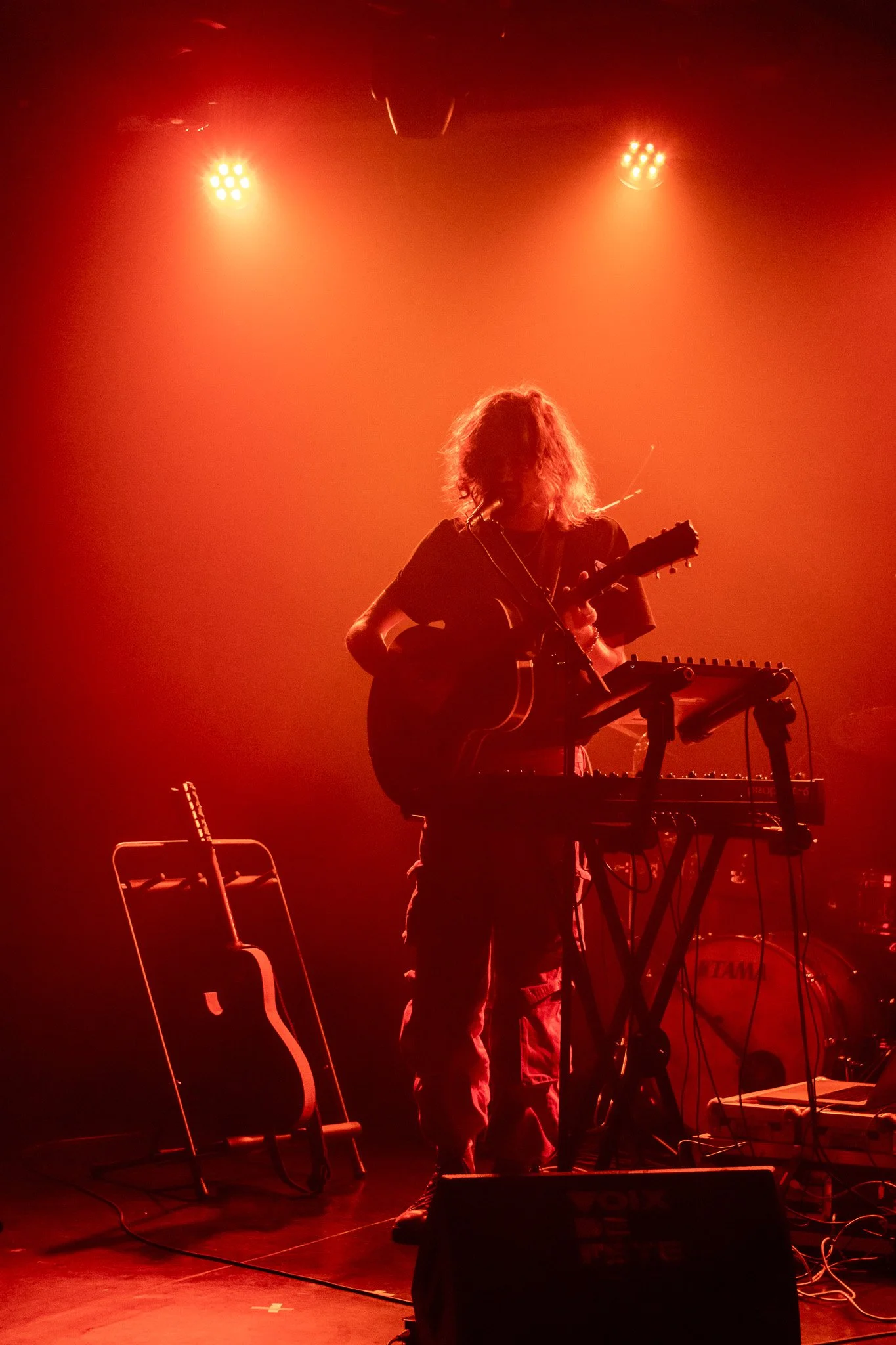 Musicien jouant de la guitare et de l'ordinateur sur scène, éclairé par des lumières rouges.