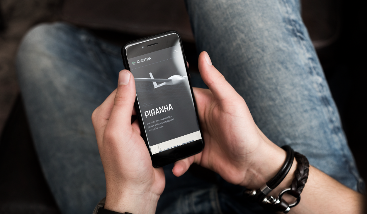Person sitting and holding a smartphone with an airplane graphic and text about the 'Piranha' drone, in a casual setting.