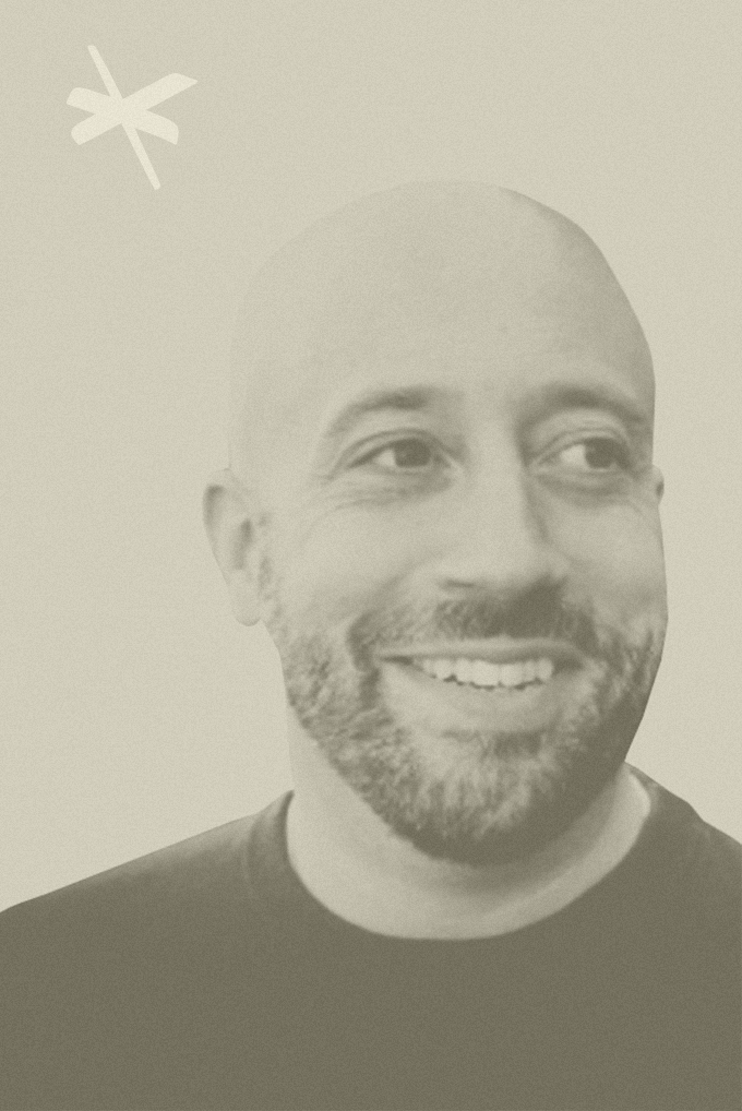 A smiling man with a beard and shaved head looking to the right, with a light background and a small star shape in the top left corner.