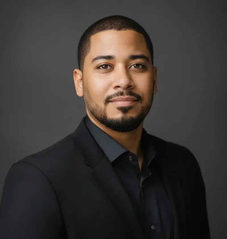 A man with short dark hair, a beard, and mustache, wearing a black blazer and shirt, against a gray background.
