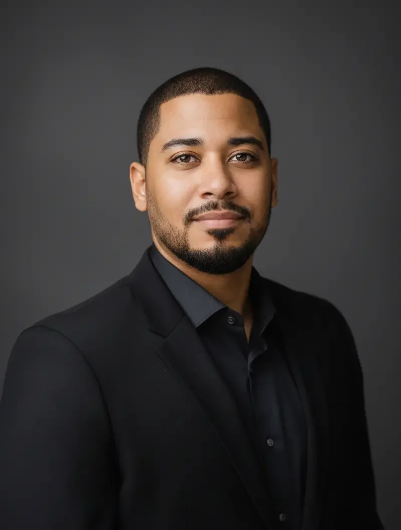 Professional headshot of a man with short dark hair, a beard, and mustache, wearing a black suit and black shirt against a dark gray background.