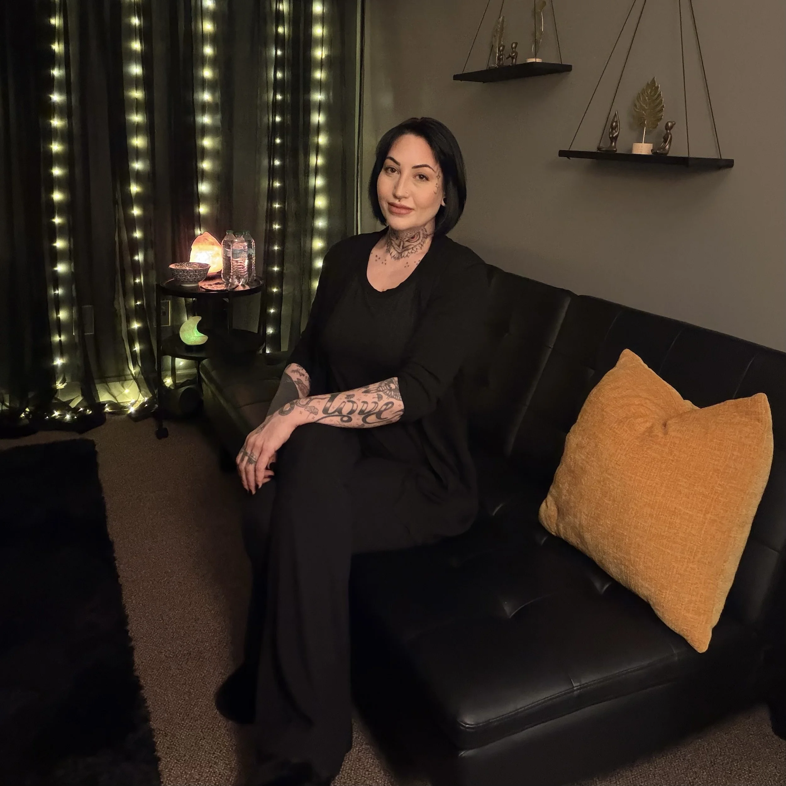 A woman with tattoos and dark hair sitting on a black couch in a dimly lit room with string lights, salt lamp, and decorative shelves on the wall.